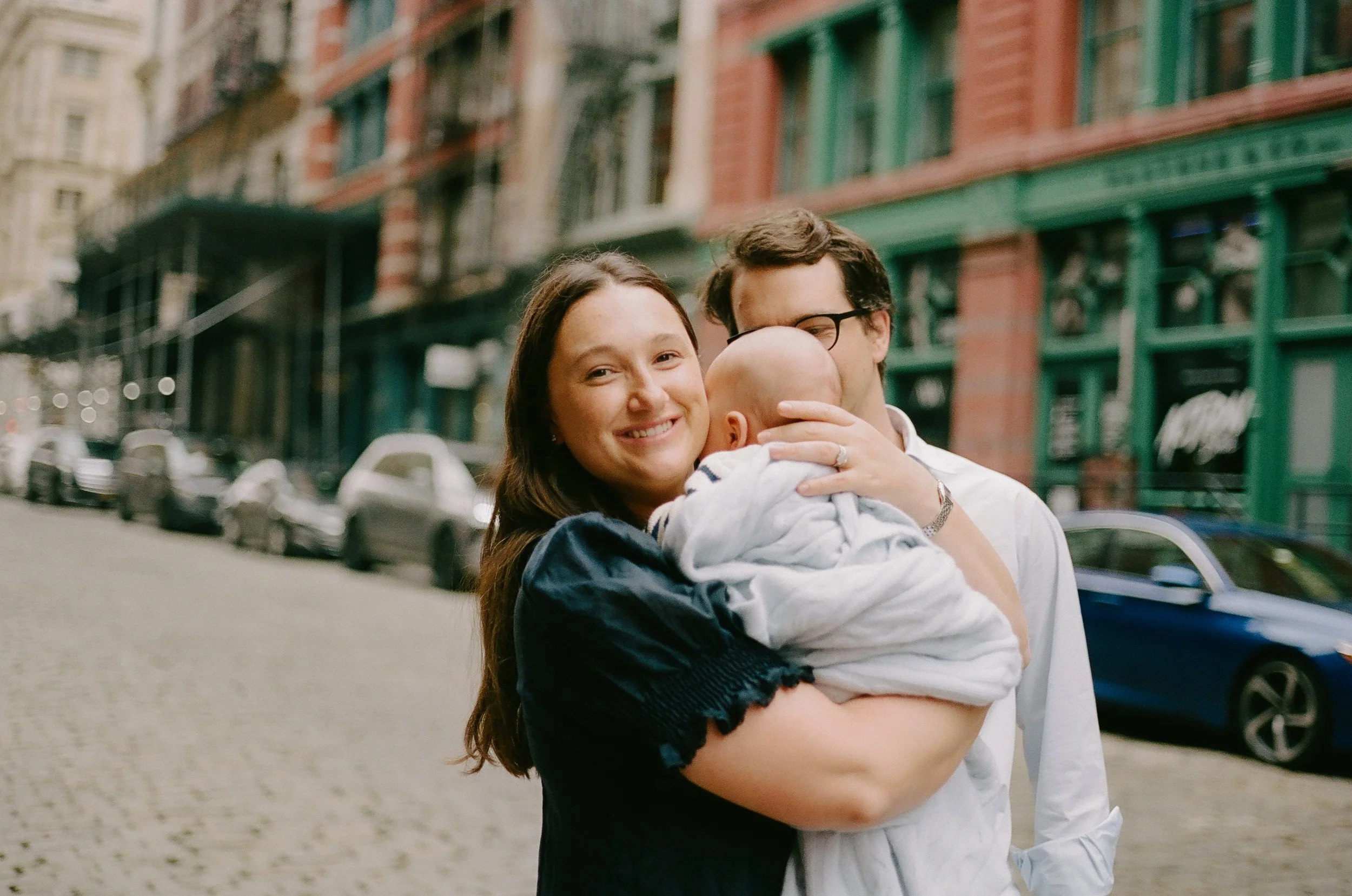 A woman holding a baby, with man in glasses hugging them, on a city street with parked cars and brick buildings in the background.