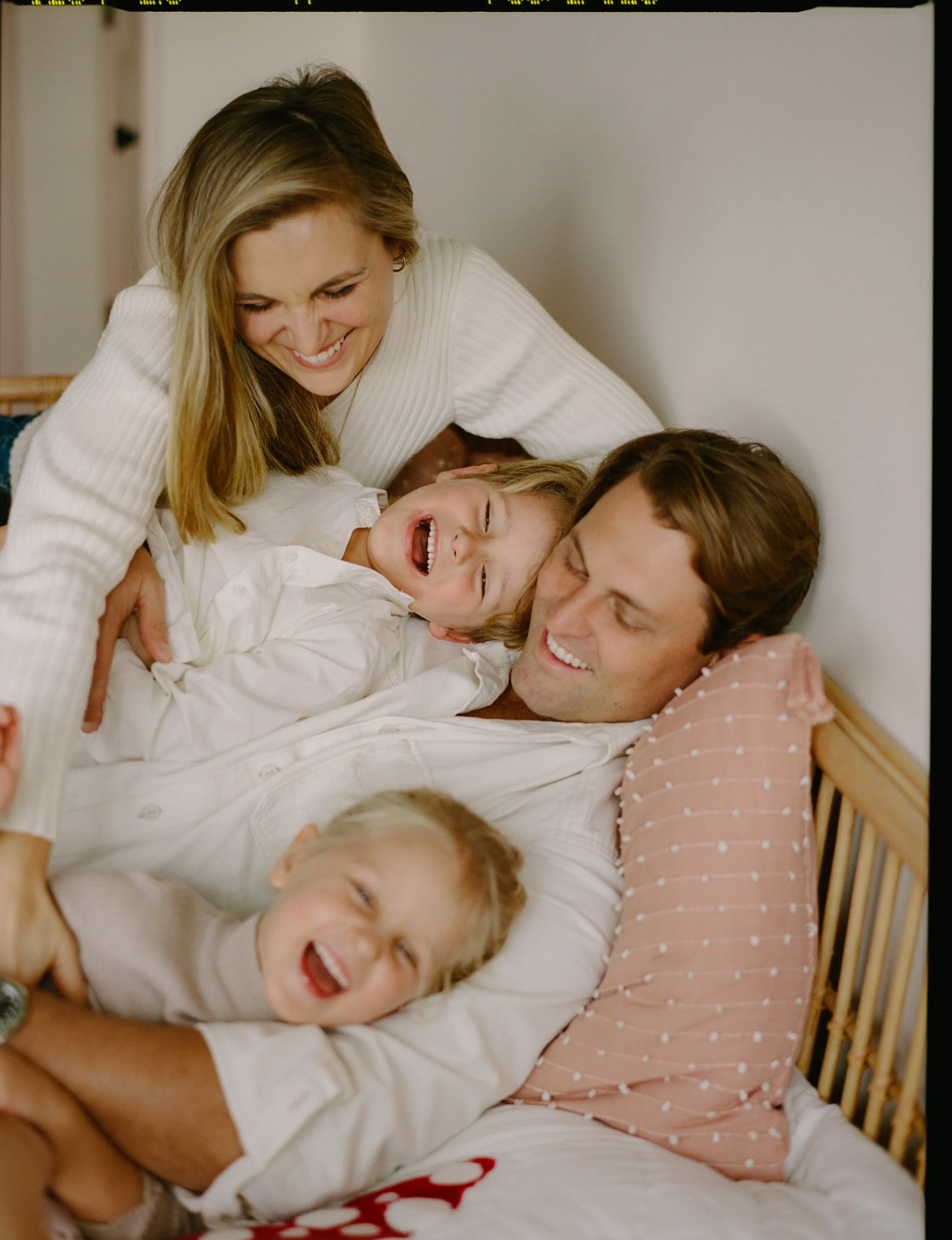 A woman, two children, and a baby are laughing and cuddling on a bed. The woman and the children are lying on their sides, facing each other, and appear to be enjoying a joyful moment together.