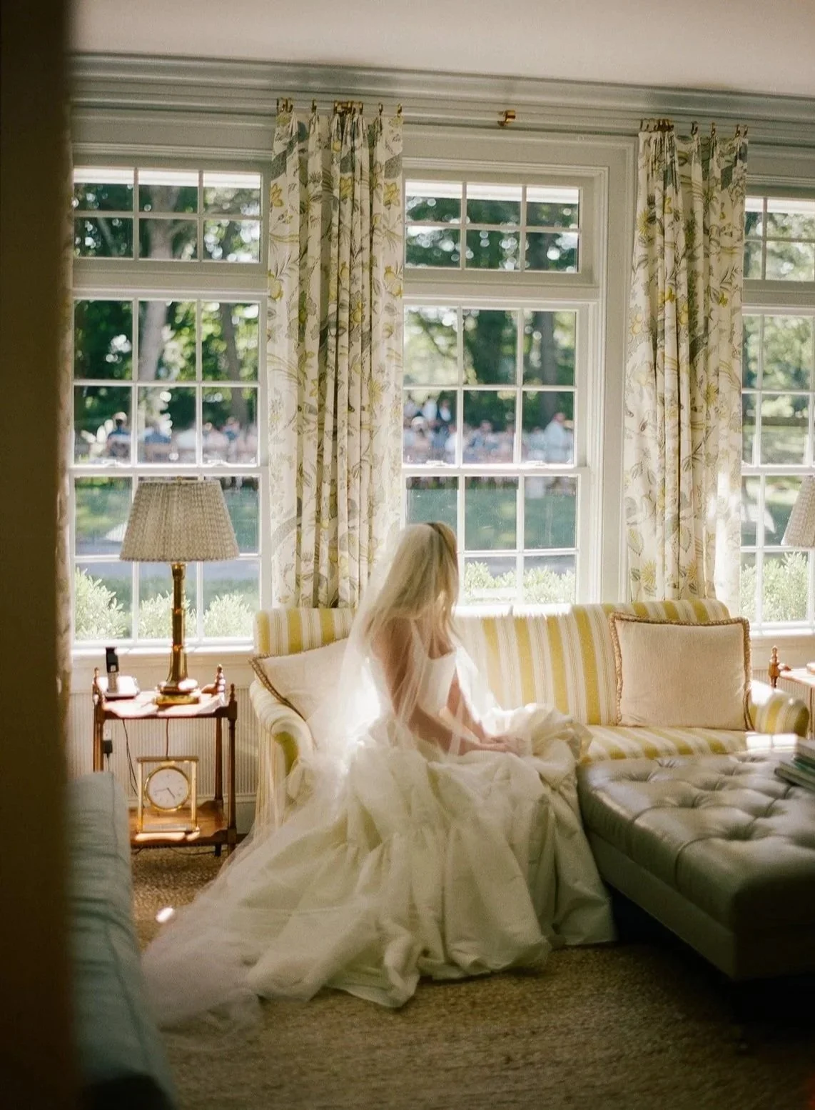 A bride sitting on a striped yellow and white sofa in a bright room with large windows and floral curtains, with her back to the camera, sunlight streaming in. Shot on medium format, contax 645