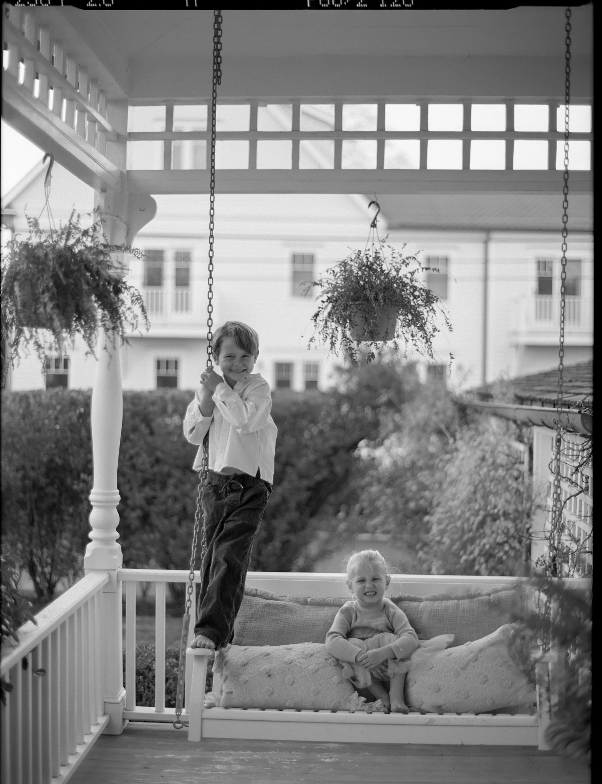 Two children on a porch with a swing. The boy stands on the swing's edge smiling, while the girl sits on the cushion of the swing, smiling and looking at the camera. There are hanging potted plants and a white railing on the porch.