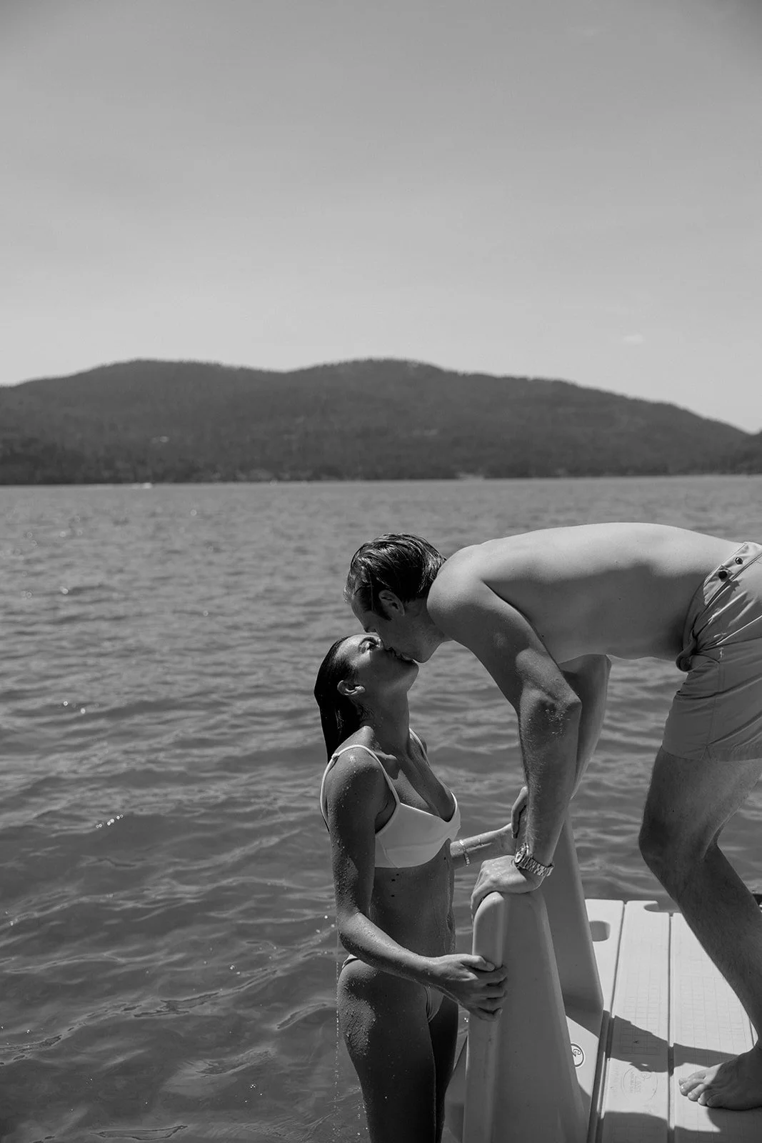 A black and white photo of a man and a woman kissing on a boat, with water and mountains in the background.