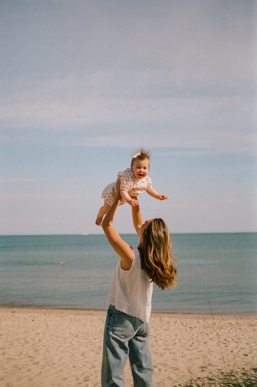 A woman lifting a happy baby girl in a dress at the beach with ocean and sky in the background.