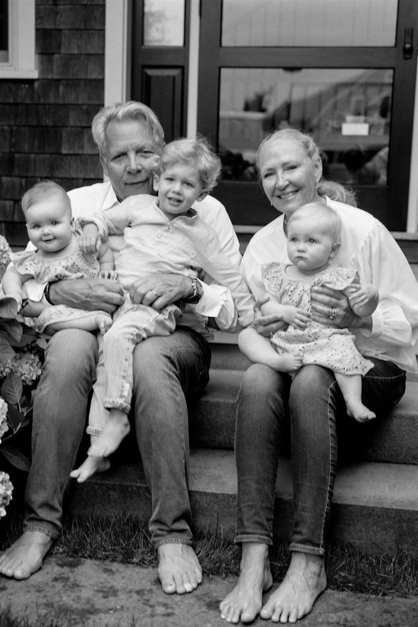 A black-and-white photo of a family sitting on a bench outside their home, including grandparents, a mother, and three young children, all with large feet and hands, smiling and looking at the camera.
