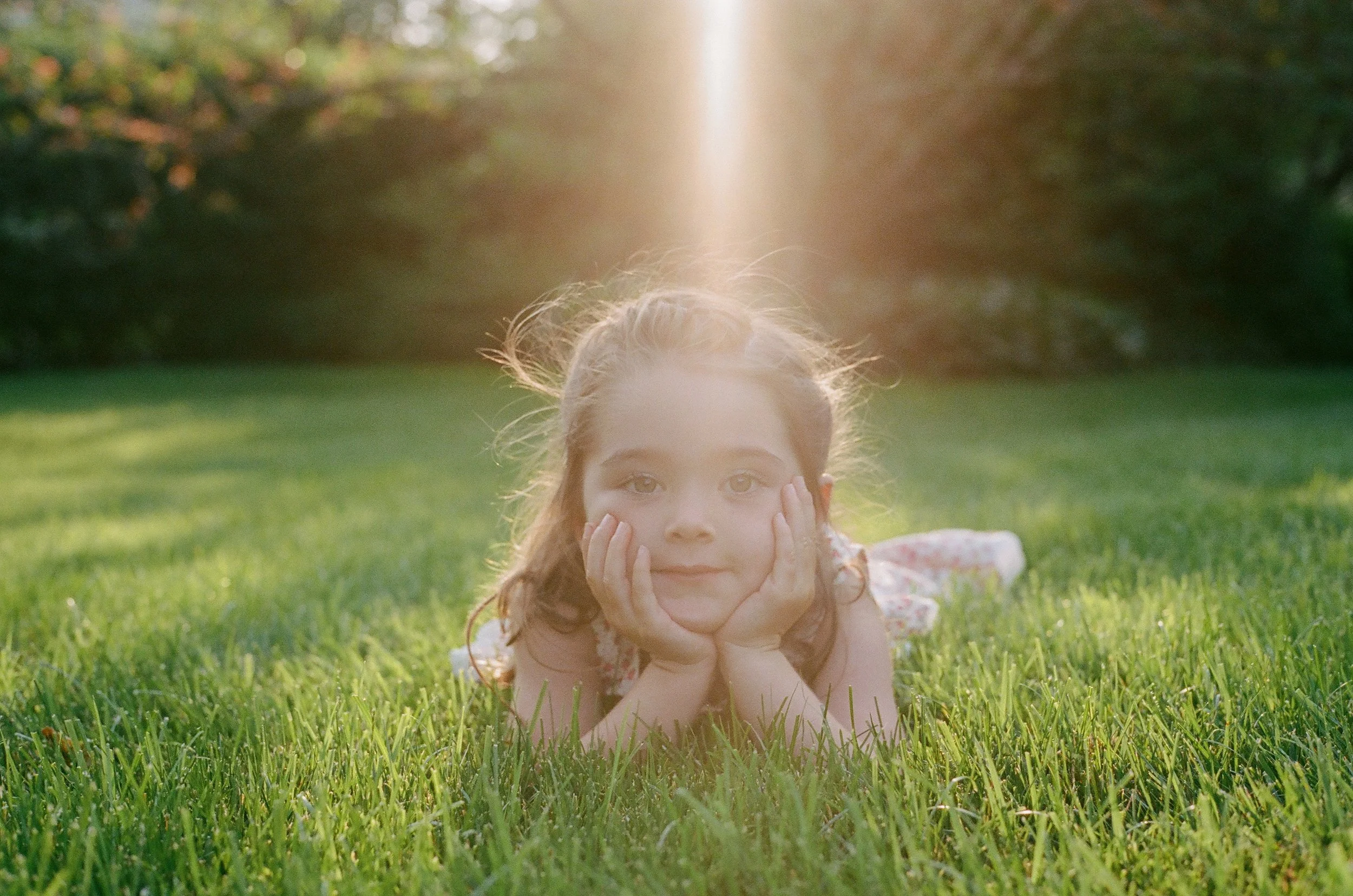 A young girl lying on her stomach on a grassy field, resting her chin in her hands, with her face close to the camera. The sunlight shines behind her, creating a warm glow and lens flare. She has light brown hair and is wearing a light-colored top.