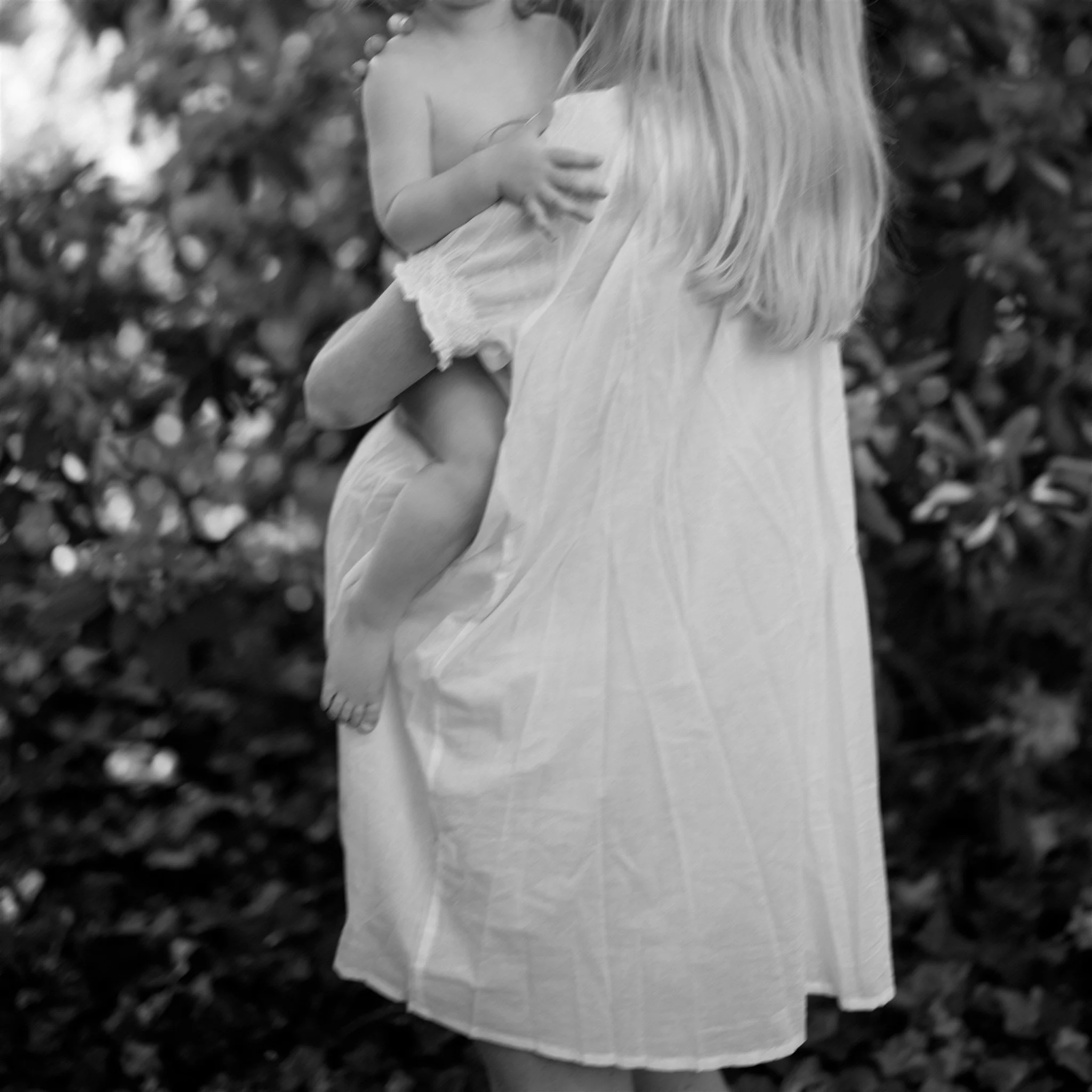 Child in a light-colored dress being held by mother, with foliage in the background.