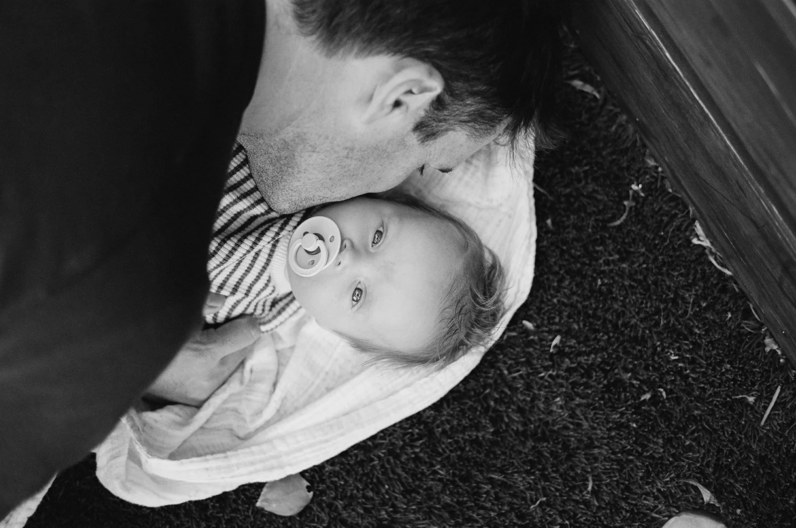 A man is lying on the ground, kissing a baby girl who has a pacifier in her mouth. The baby is lying on a blanket on a carpeted surface.