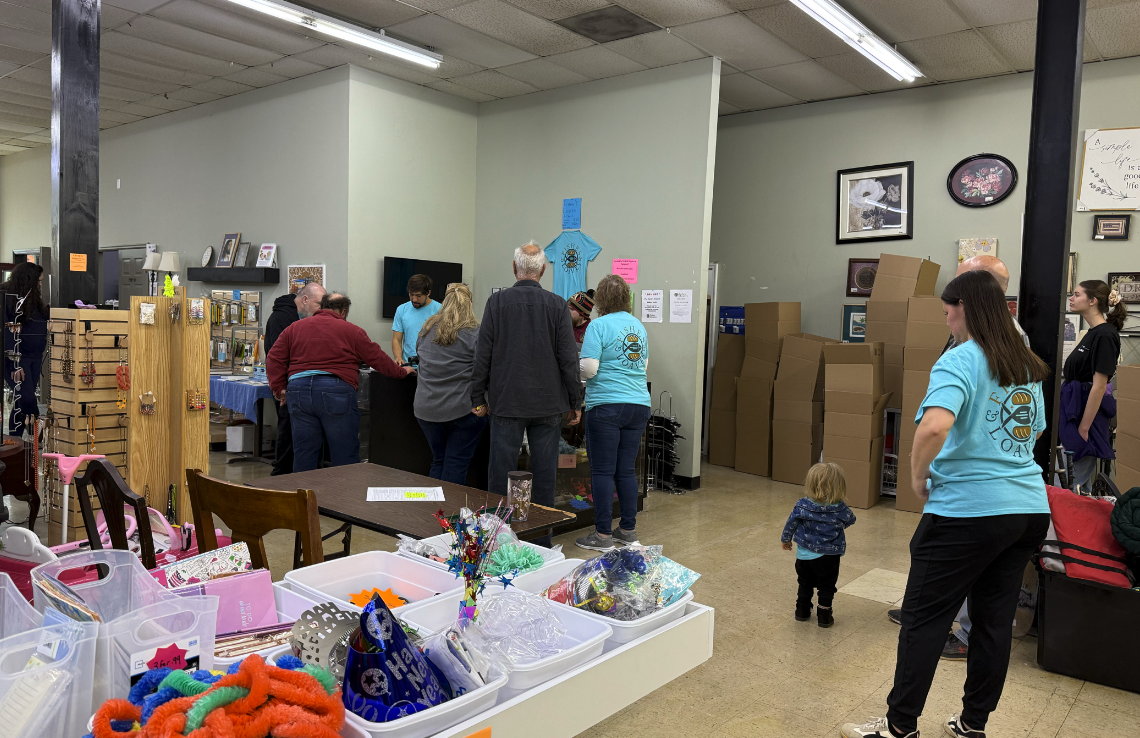 People shopping at a donation or thrift store, with some browsing items and others standing in line, surrounded by various items on tables and shelves, and a partially empty wall with framed pictures and stacked boxes in the background.