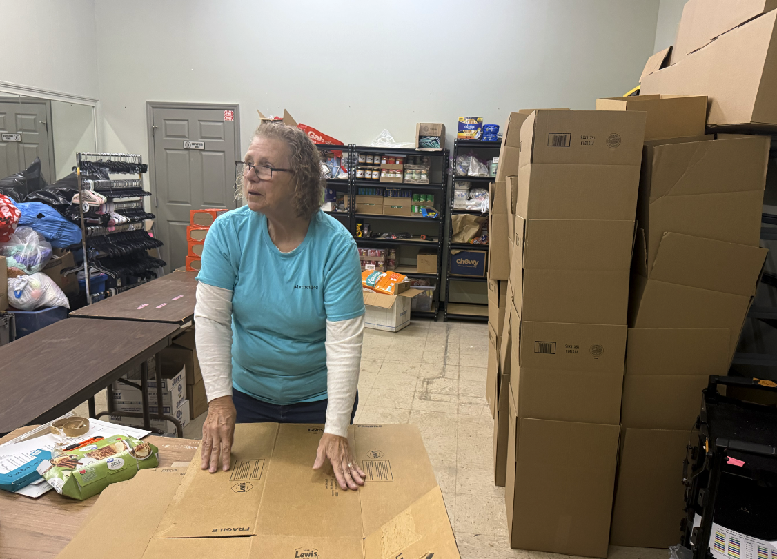 A woman wearing glasses and a light blue shirt standing behind a table in a storage room, with large stacks of cardboard boxes on her right and shelves stocked with various supplies in the background.