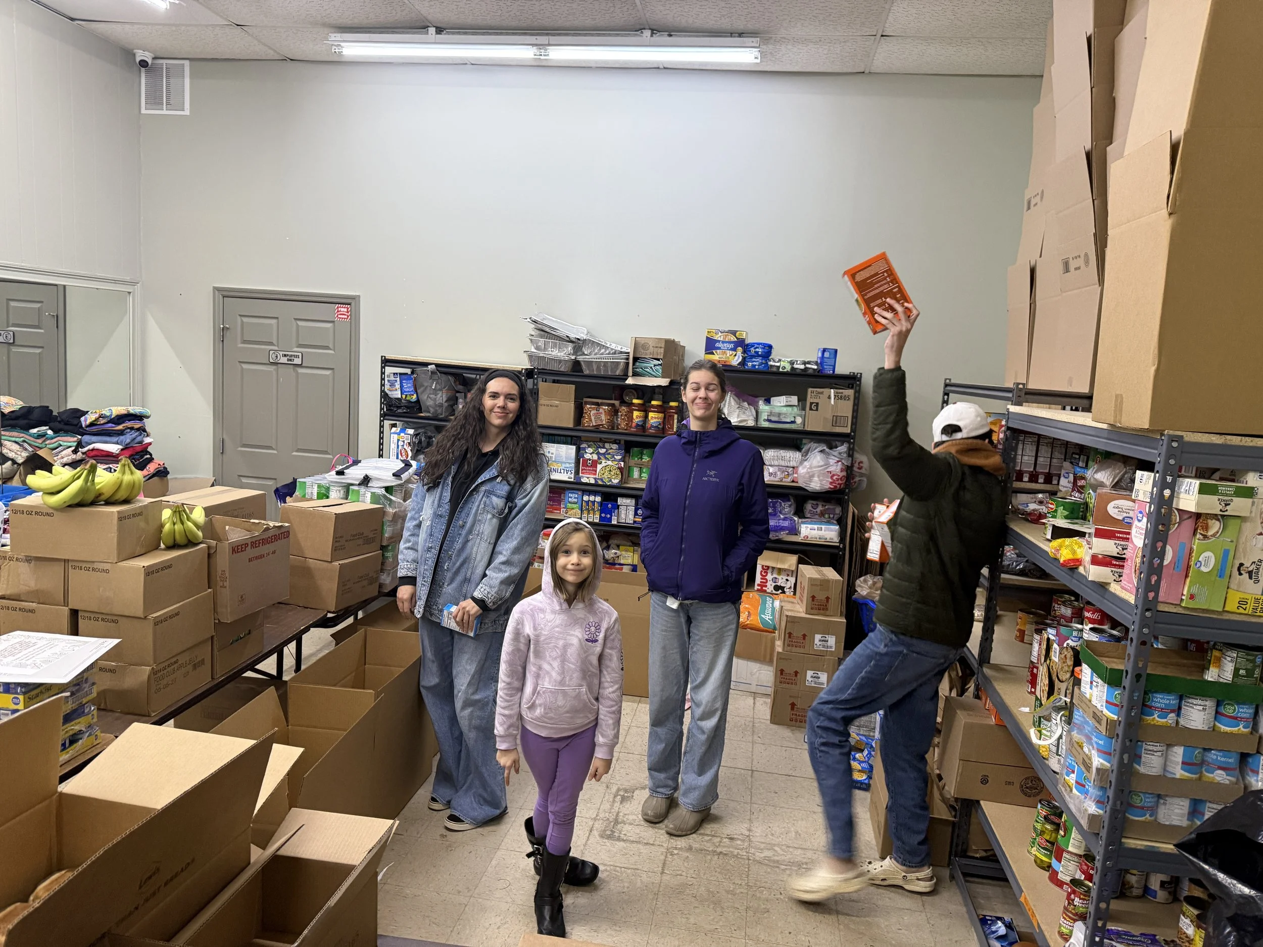 People shopping at a food bank or pantry arranged with cardboard boxes, shelves stocked with canned goods, and a table with bananas.