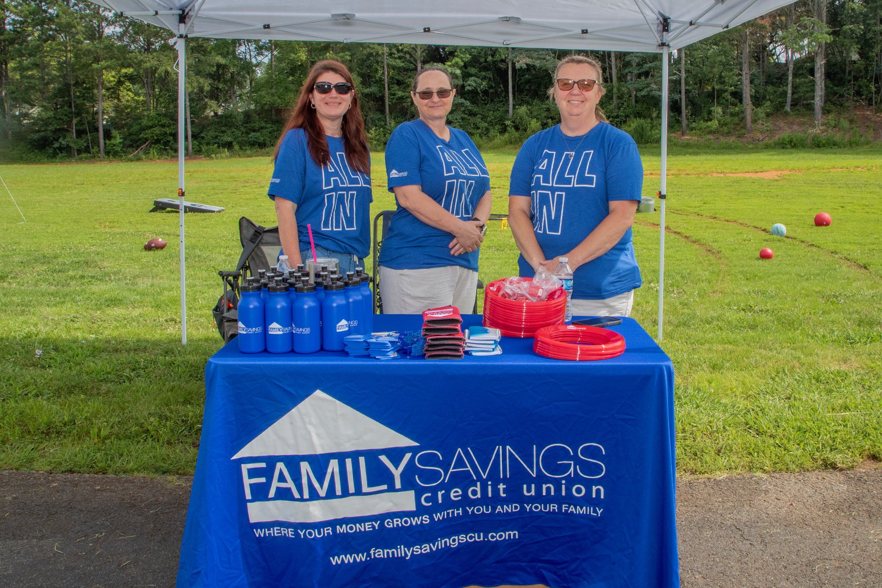 Three women standing behind a table with promotional materials at an outdoor event, wearing matching blue t-shirts with 'ALL IN' printed on them, under a white canopy on a grassy field with trees in the background.