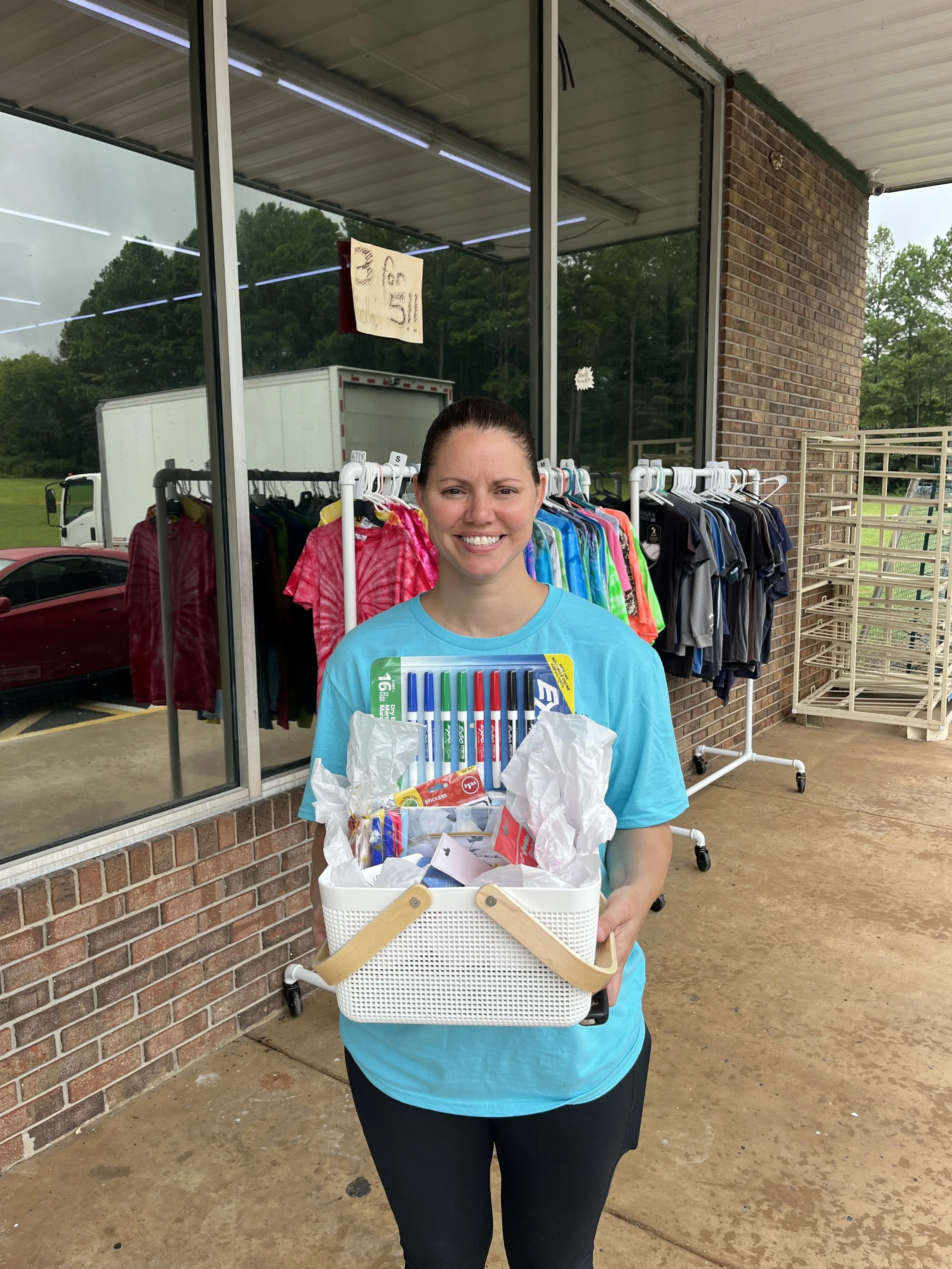 Woman holding a white basket filled with pens, stickers, and various small items outside a store with clothing on racks behind her.