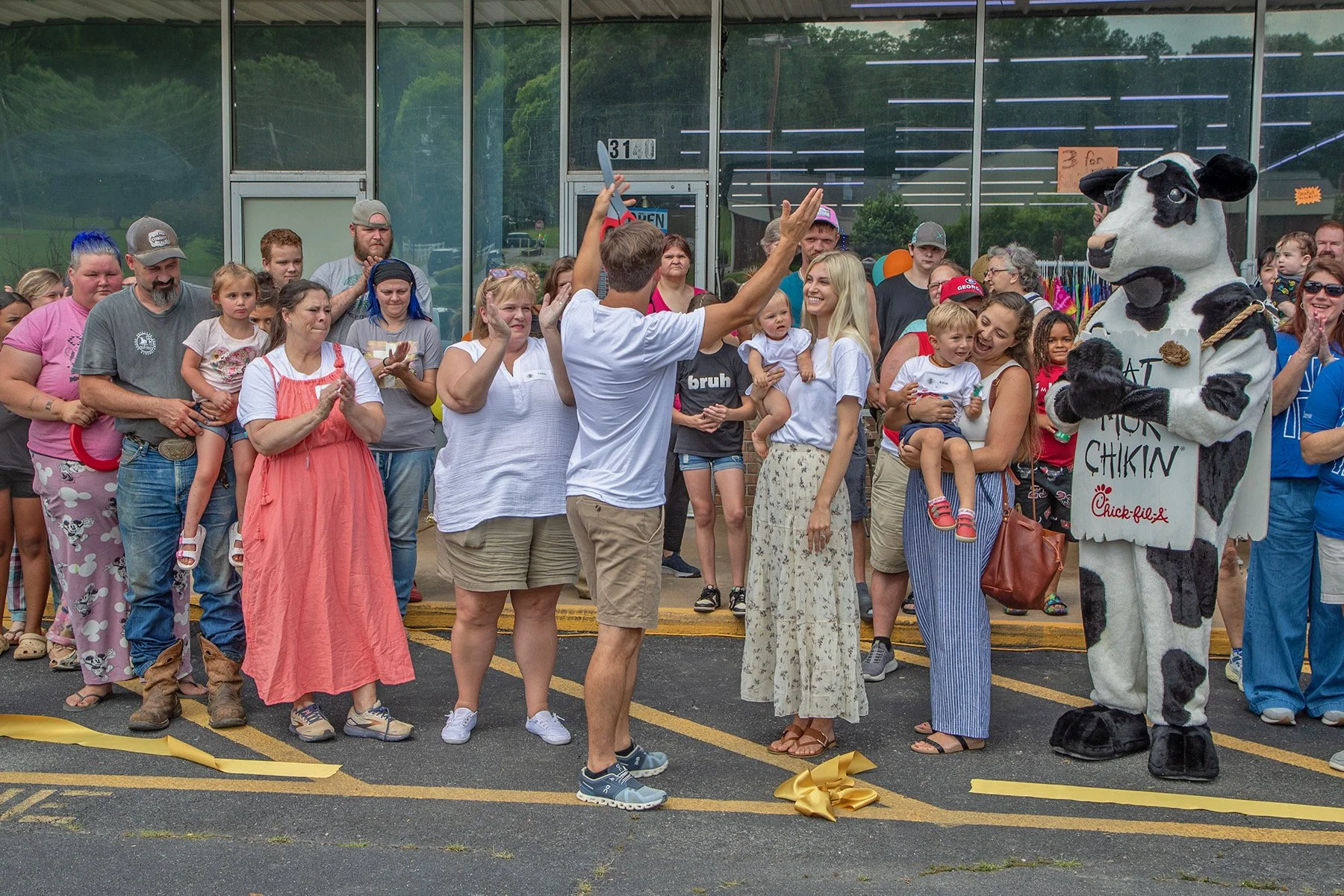 A crowd of people gathered outside a store during a ribbon-cutting event, including a person in a cow costume holding a sign that reads 'Eat Mor Chikin', with children and adults smiling and clapping.