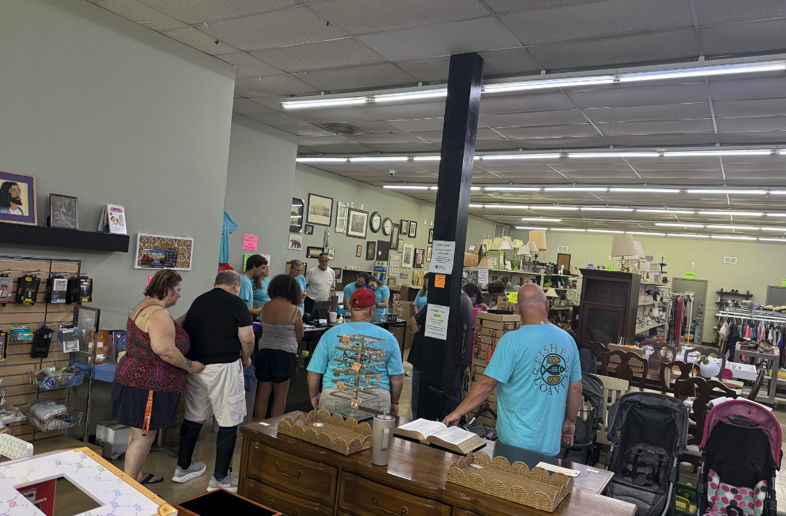People shopping and inspecting items at a thrift store, with shelves displaying various goods, framed pictures on the wall, and a group of people gathered around a counter.