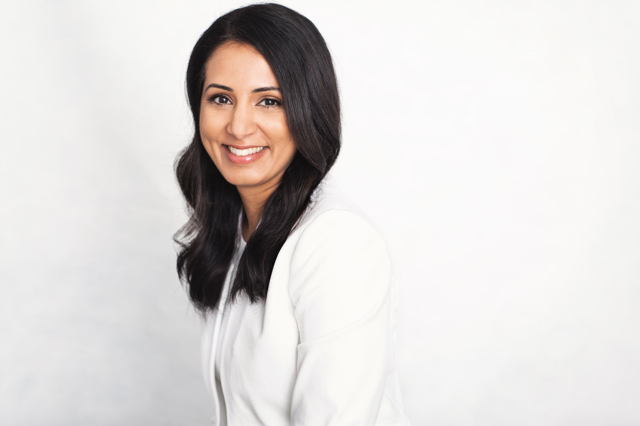 A woman with long black hair smiling, wearing a white blazer, against a plain white background.
