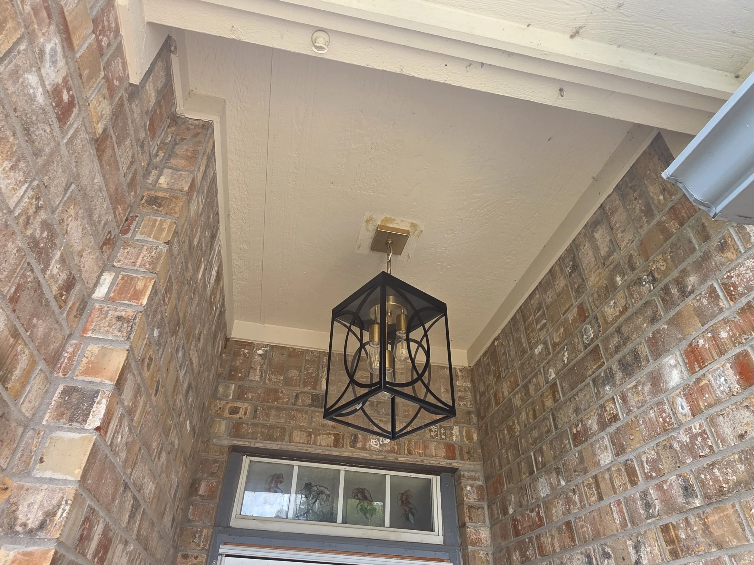 A black metal lantern-style chandelier hanging from a ceiling with a textured beige surface, surrounded by brick wall corners and a window with stickers of flowers and cartoon characters.
