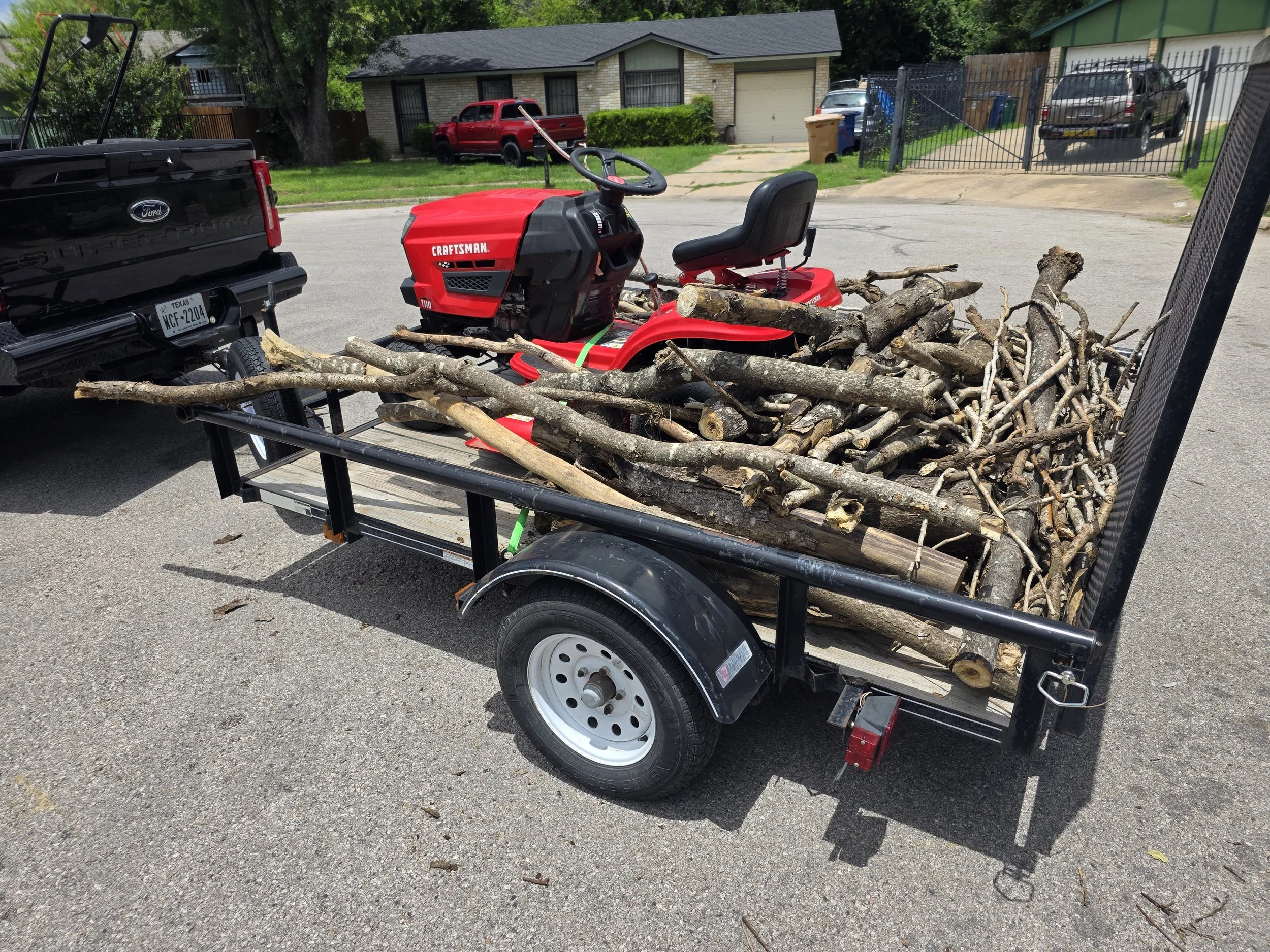 A trailer carrying a pile of cut tree branches and logs parked on a street in front of residential houses.
