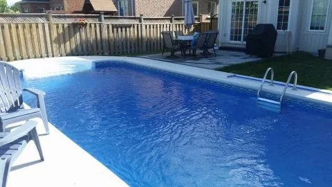 Backyard swimming pool with a deck, outdoor dining table, and chairs, enclosed by a wooden fence.