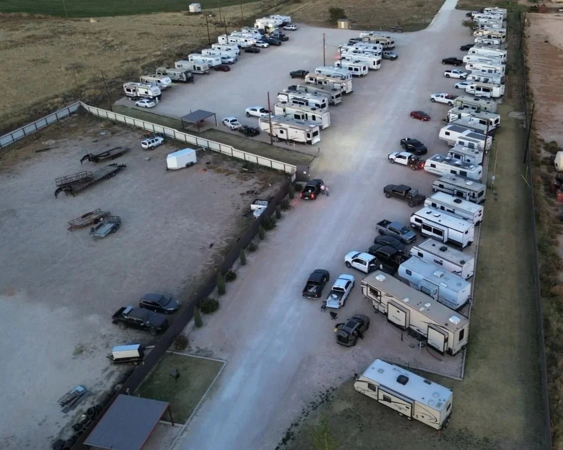 Aerial view of a parking lot filled with numerous RVs and trailers, with some cars parked around them. The lot is enclosed by a fence, with open land surrounding it.