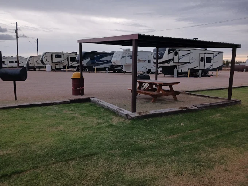 A covered picnic area with a wooden picnic table, situated on a concrete slab, next to a grassy lawn area. In the background, several travel trailers or RVs are parked in an open lot.