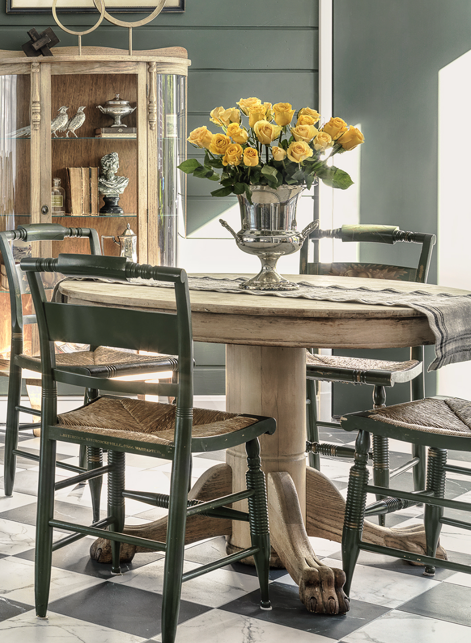 A dining room with a round wooden table, a large silver vase with yellow roses, four black chairs with woven seats, a green shiplap wall, and a wooden display cabinet with decorative objects and books.
