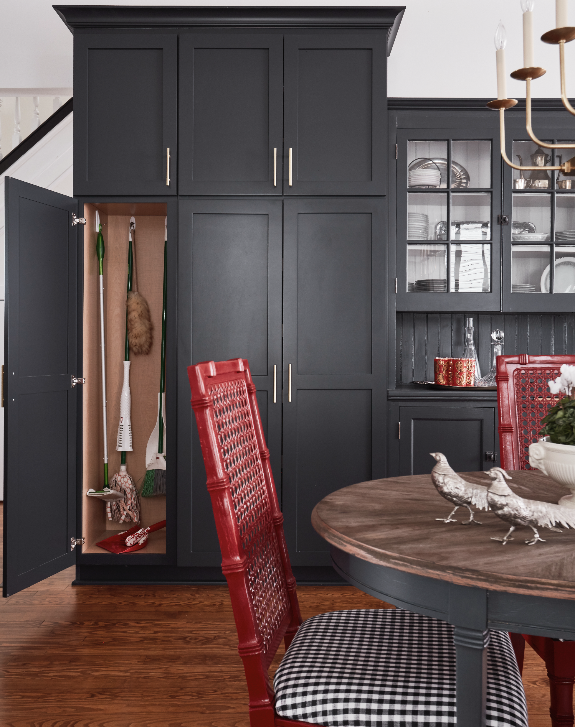 A dark blue kitchen cabinet with a glass-paneled door, displaying plates and bowls inside. An open pantry contains cleaning supplies and a dustpan with a fur broom. A round wooden table with a checkered tablecloth and decorative bird figurines is in the foreground, and red dining chairs surround the table. The room has a hardwood floor.