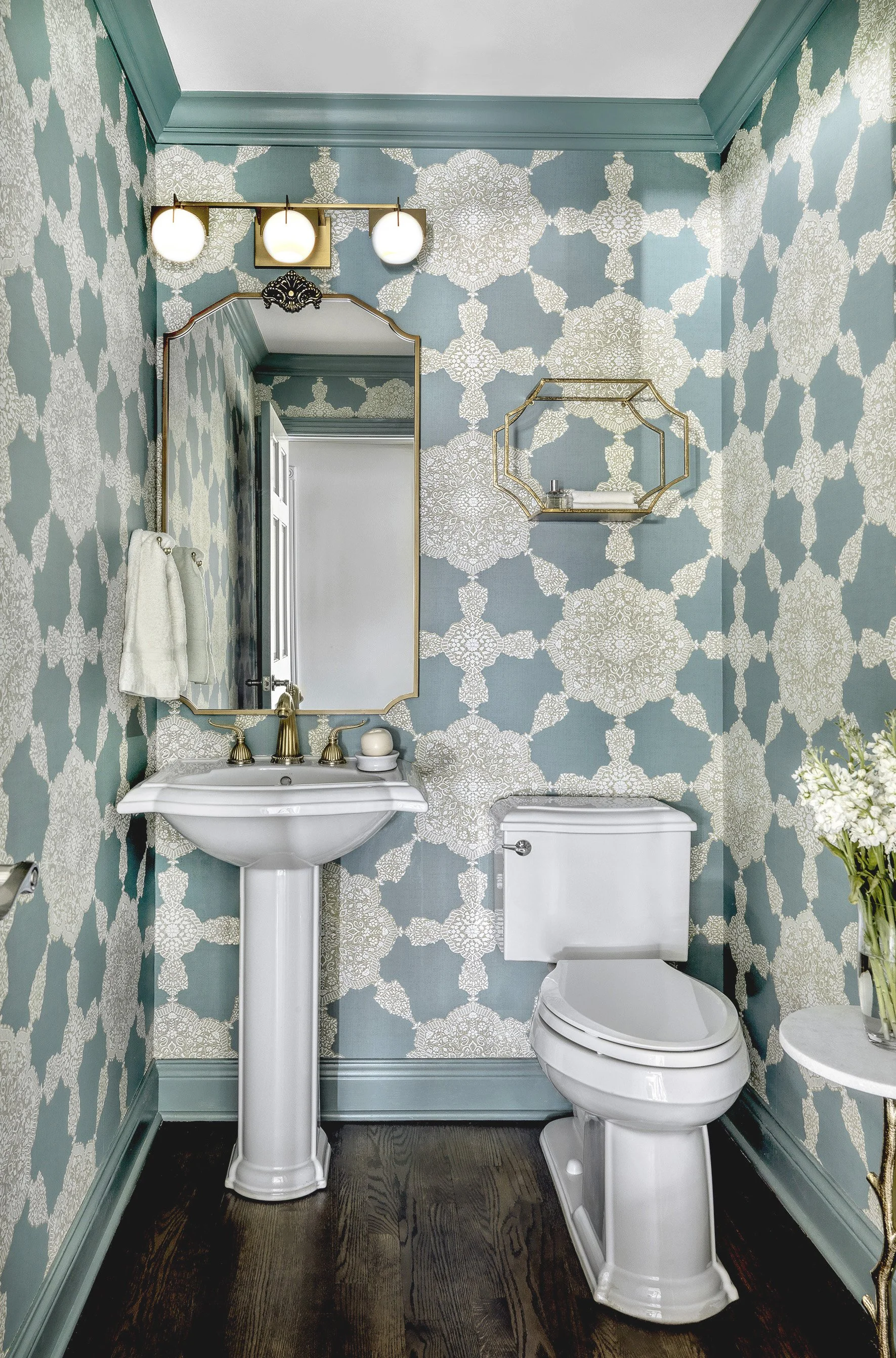 A bathroom with teal decorative patterned wallpaper, gold accents, a white pedestal sink, a toilet and a mirror.