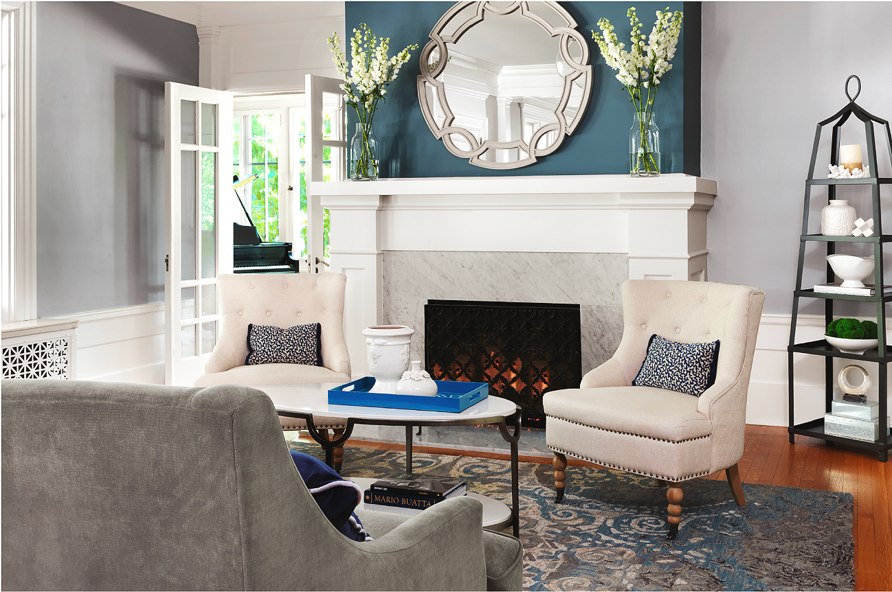 Living room with white fireplace, large mirror, two armchairs with pillows, a coffee table with decorative items, a patterned rug, and a black shelf with decor.