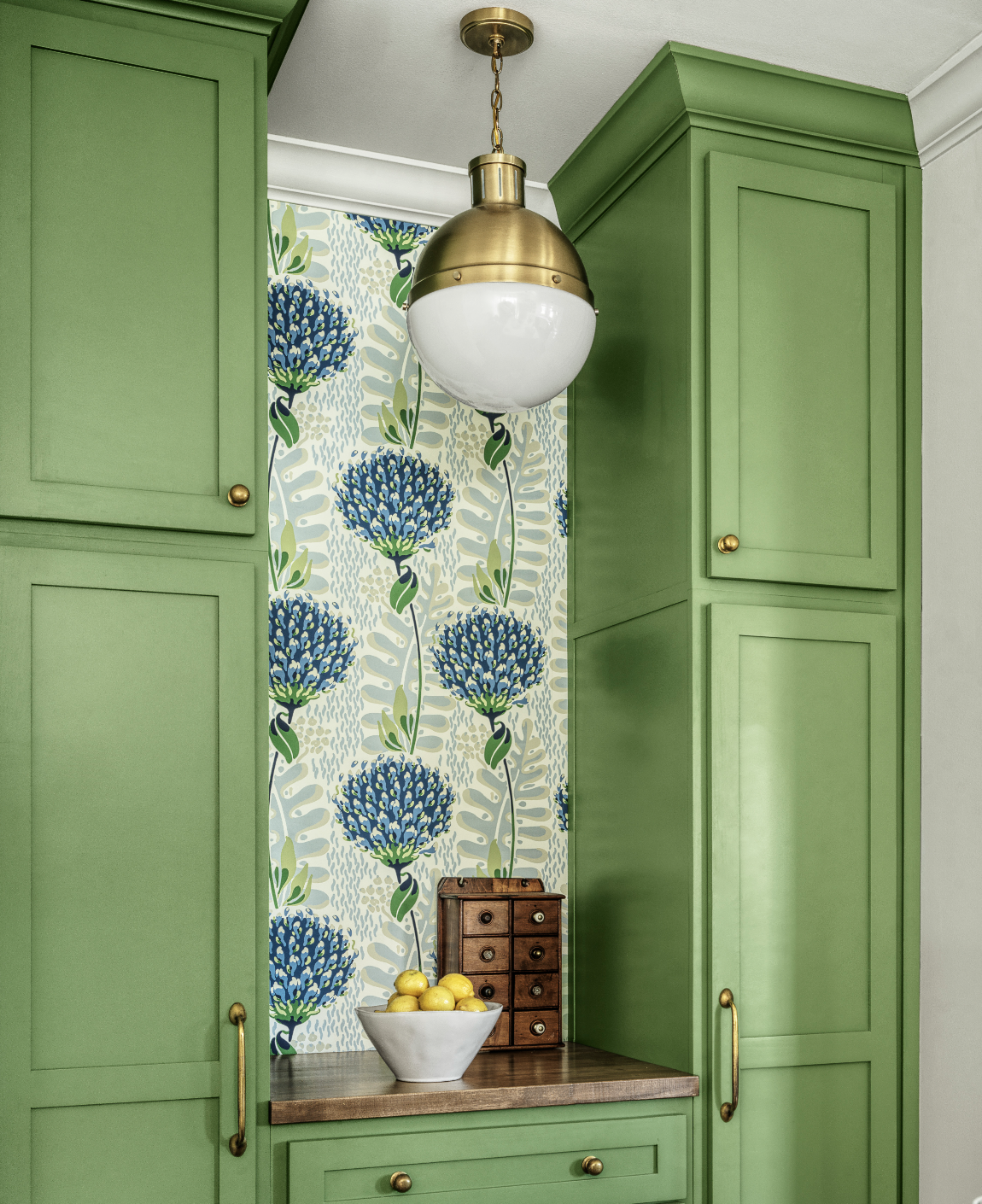 A kitchen corner with green cabinets and a floral wallpaper with blue and green flowers. There is a brass and white globe pendant light, a wooden countertop with a white bowl of yellow lemons, and a small wooden storage box with drawers.