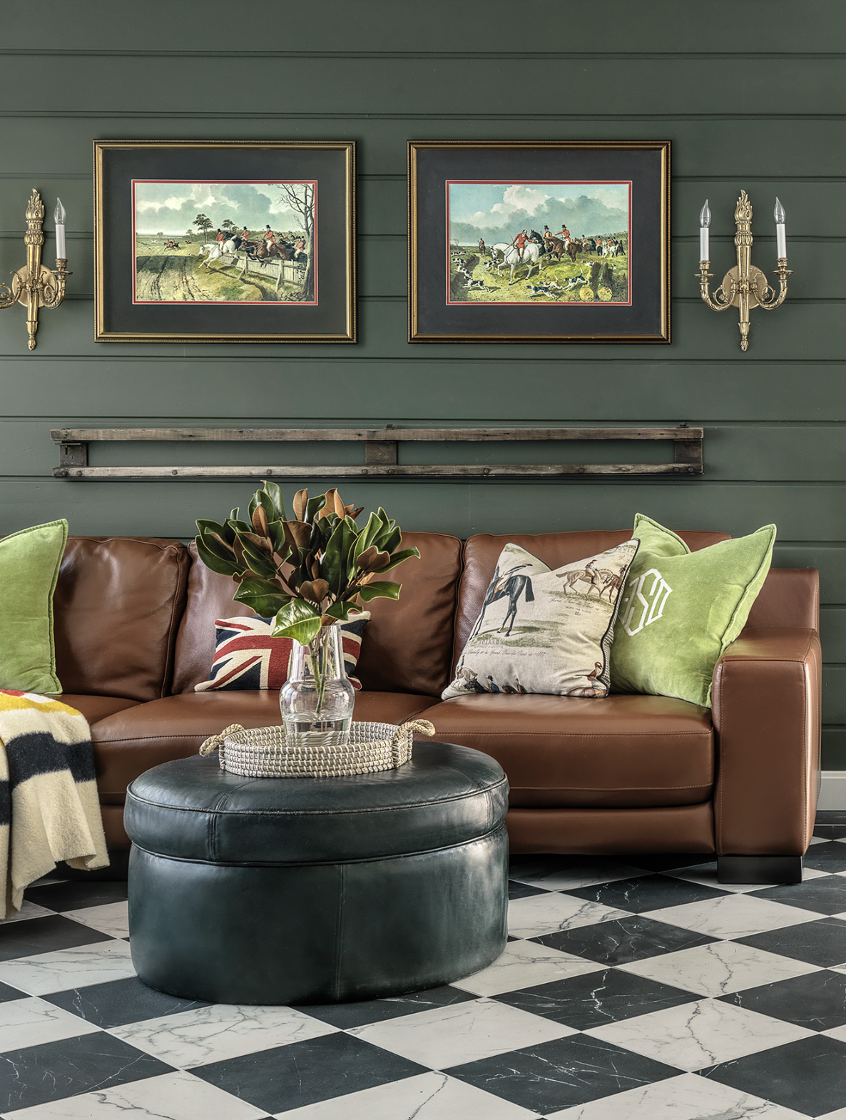 Living room with a brown leather sofa decorated with green and patterned pillows, a glass vase with a plant on an ottoman, black and white checkered flooring, green shiplap walls with framed horse racing images, and gold wall sconces.