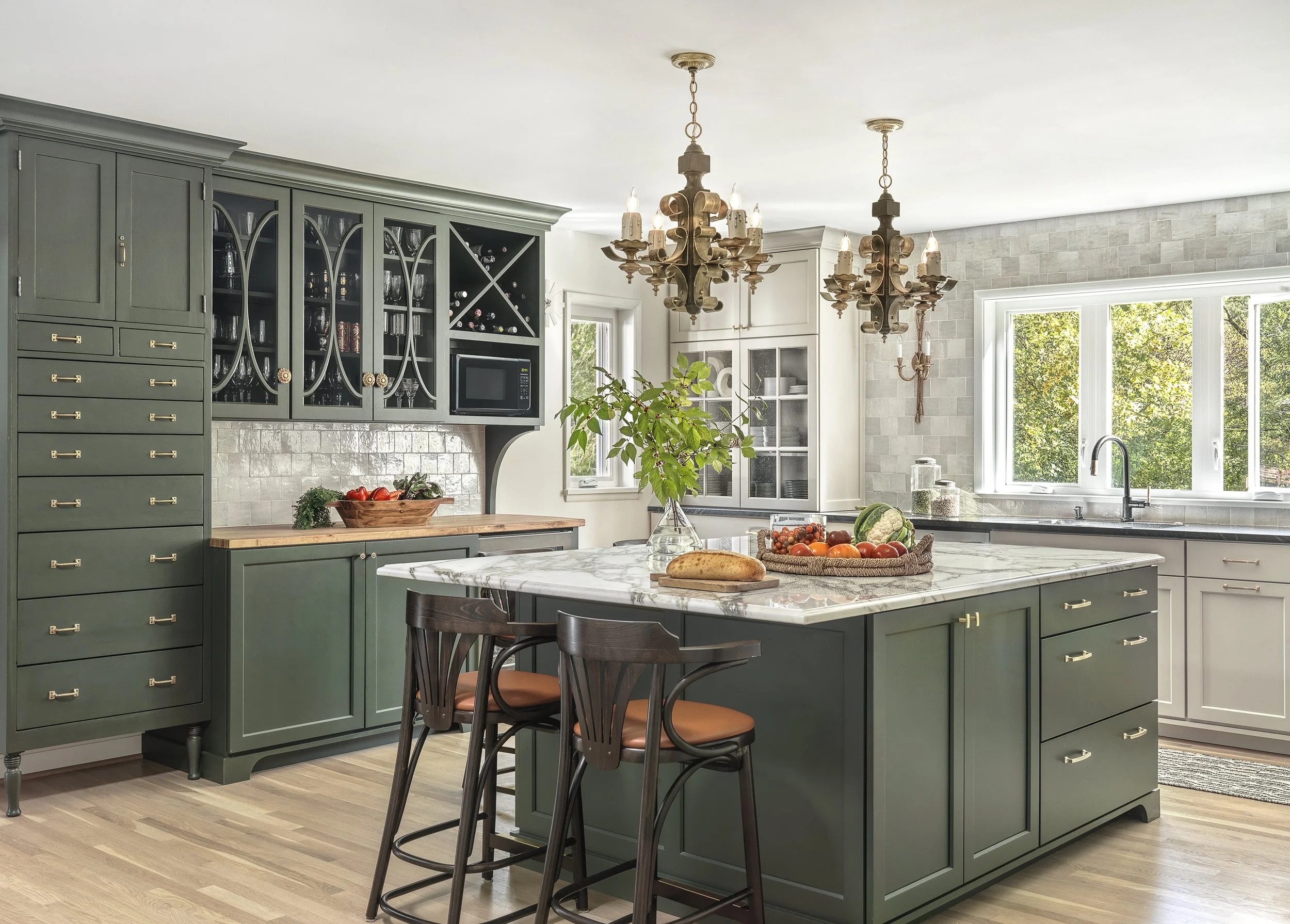 A modern kitchen with dark green cabinets, a marble island, pendant chandeliers, and a window showing trees outside.