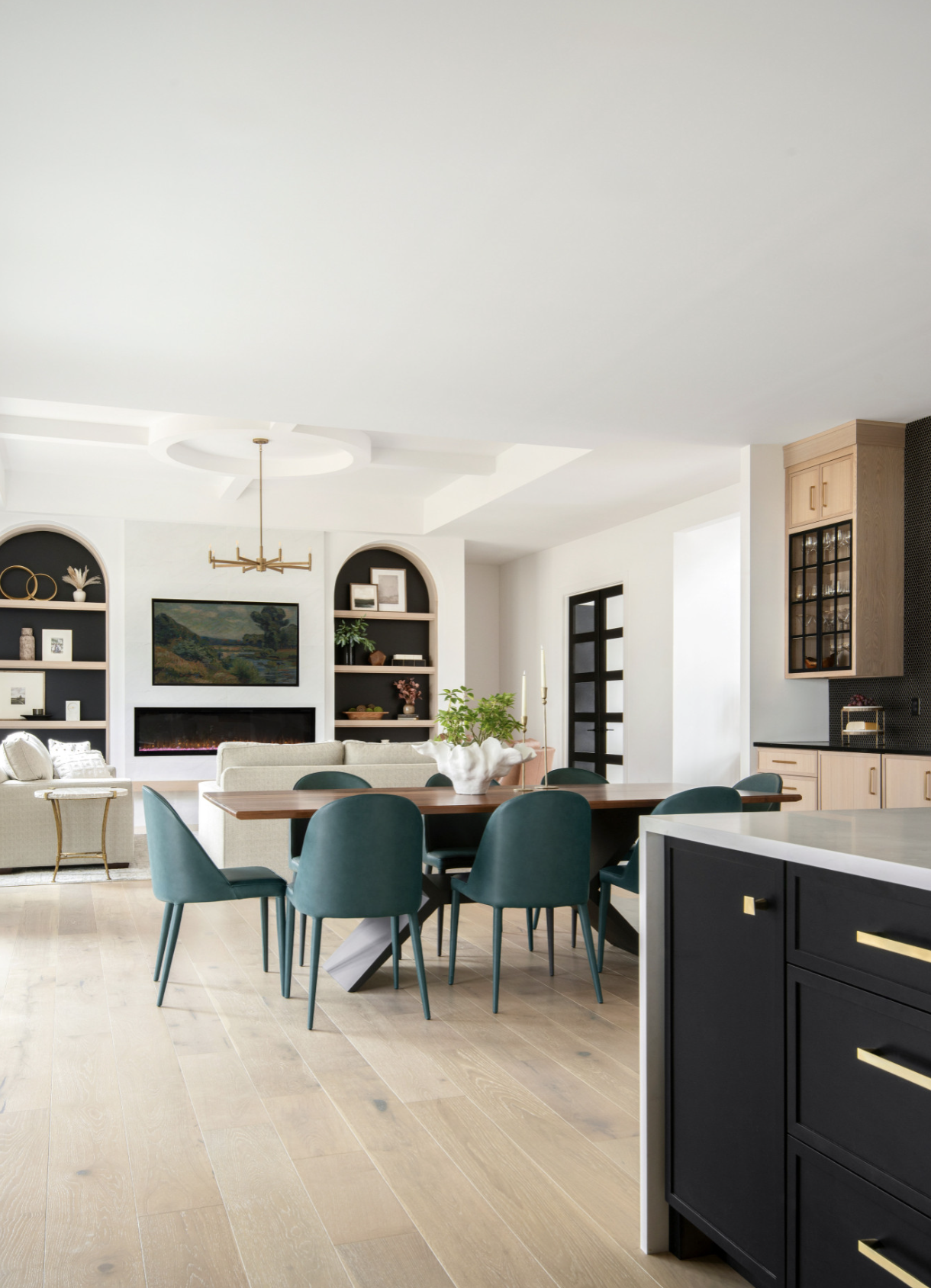 Open-concept living and dining area with a fireplace, built-in shelves, and a kitchen with black and wood cabinets, featuring a wooden dining table with teal chairs, and light hardwood floors.