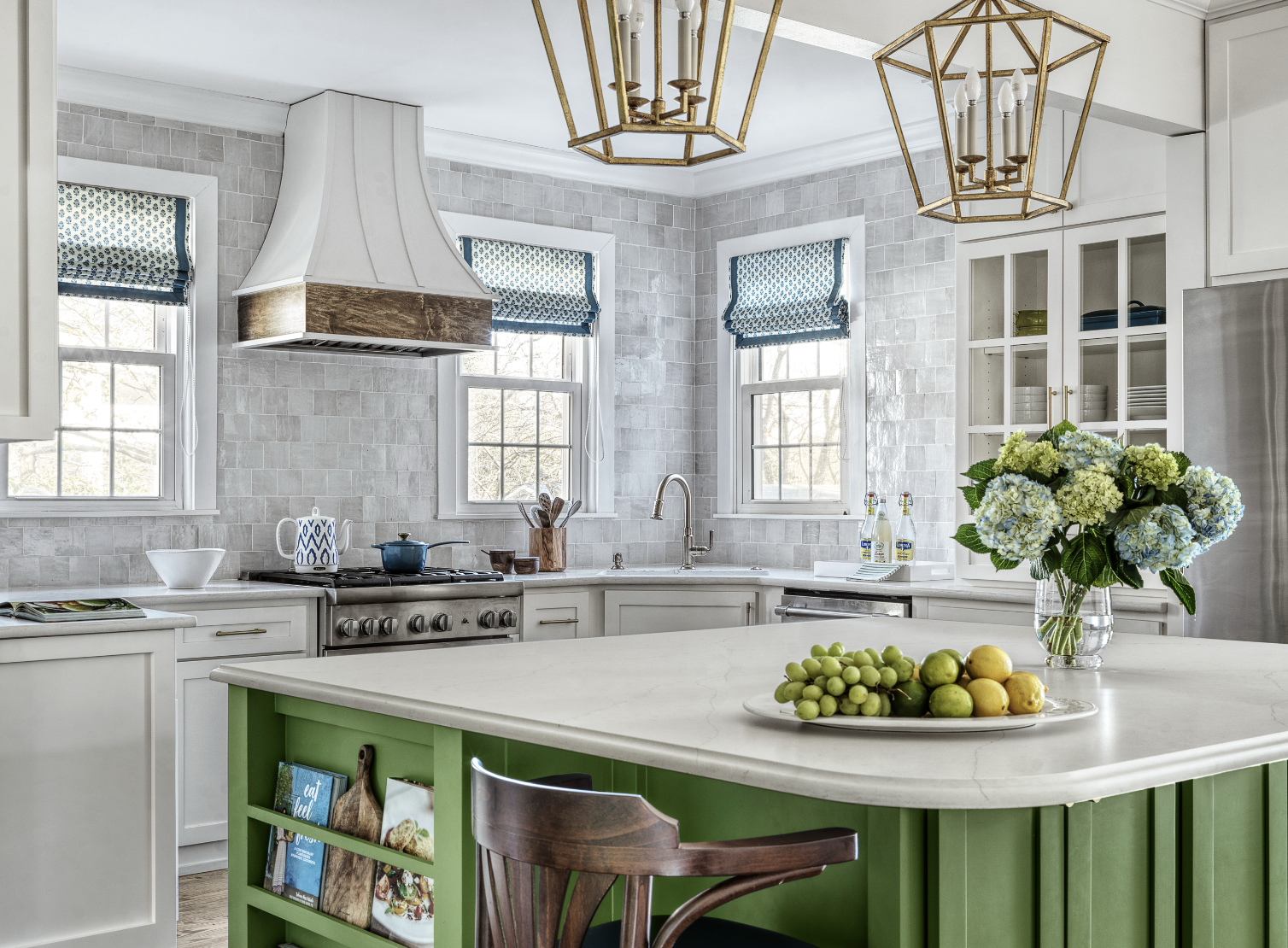 Bright kitchen with a white marble countertop island with a green base. Two brass and glass pendant lights hang above. A vase of blue and white hydrangeas and a plate of green grapes and pears are on the island. Three windows with blue patterned Roman shades, a gas stove, and a chandelier are visible.