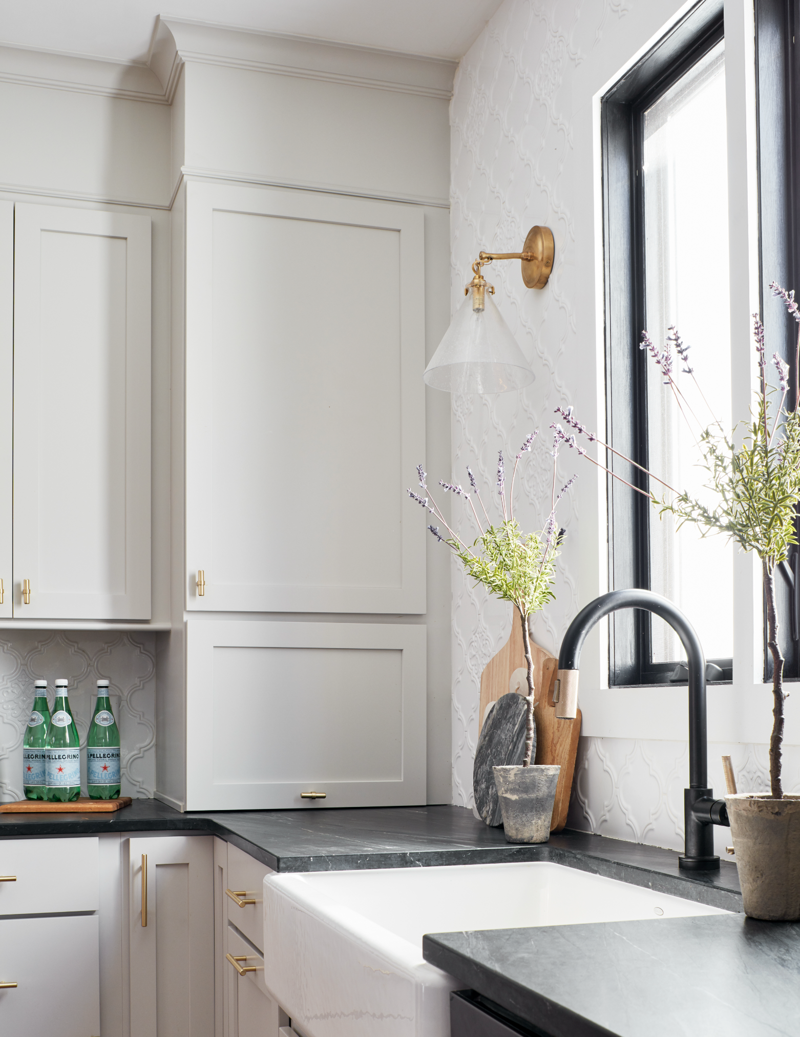 A modern kitchen with white cabinets, black countertops, and a white farmhouse sink. There is a window with a black frame letting in natural light. Decorative items include a potted plant, a cutting board, and a wall-mounted light fixture. Bottled sparkling water is on a wooden tray near the sink.