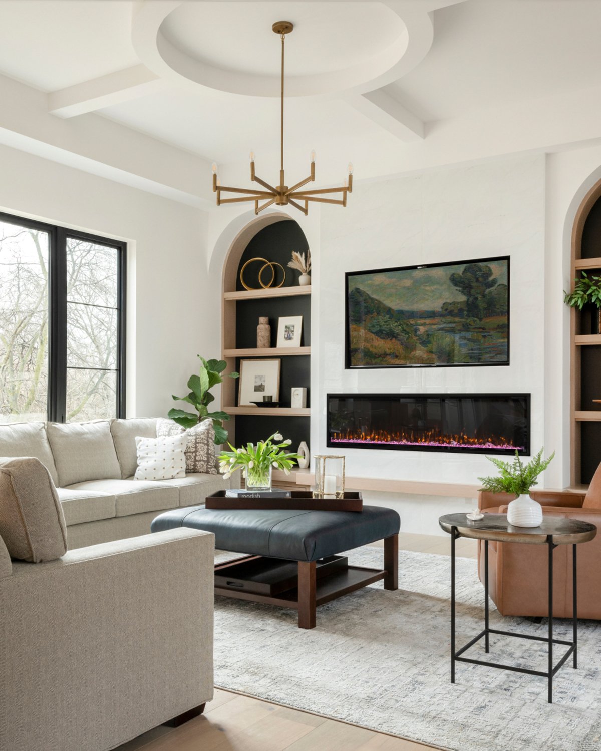 Contemporary living room with white walls, beige sectional sofa and black coffee table. Large window, modern fireplace and wall-mounted TV. Shelves with decor, minimalist chandelier, and neutral rug.