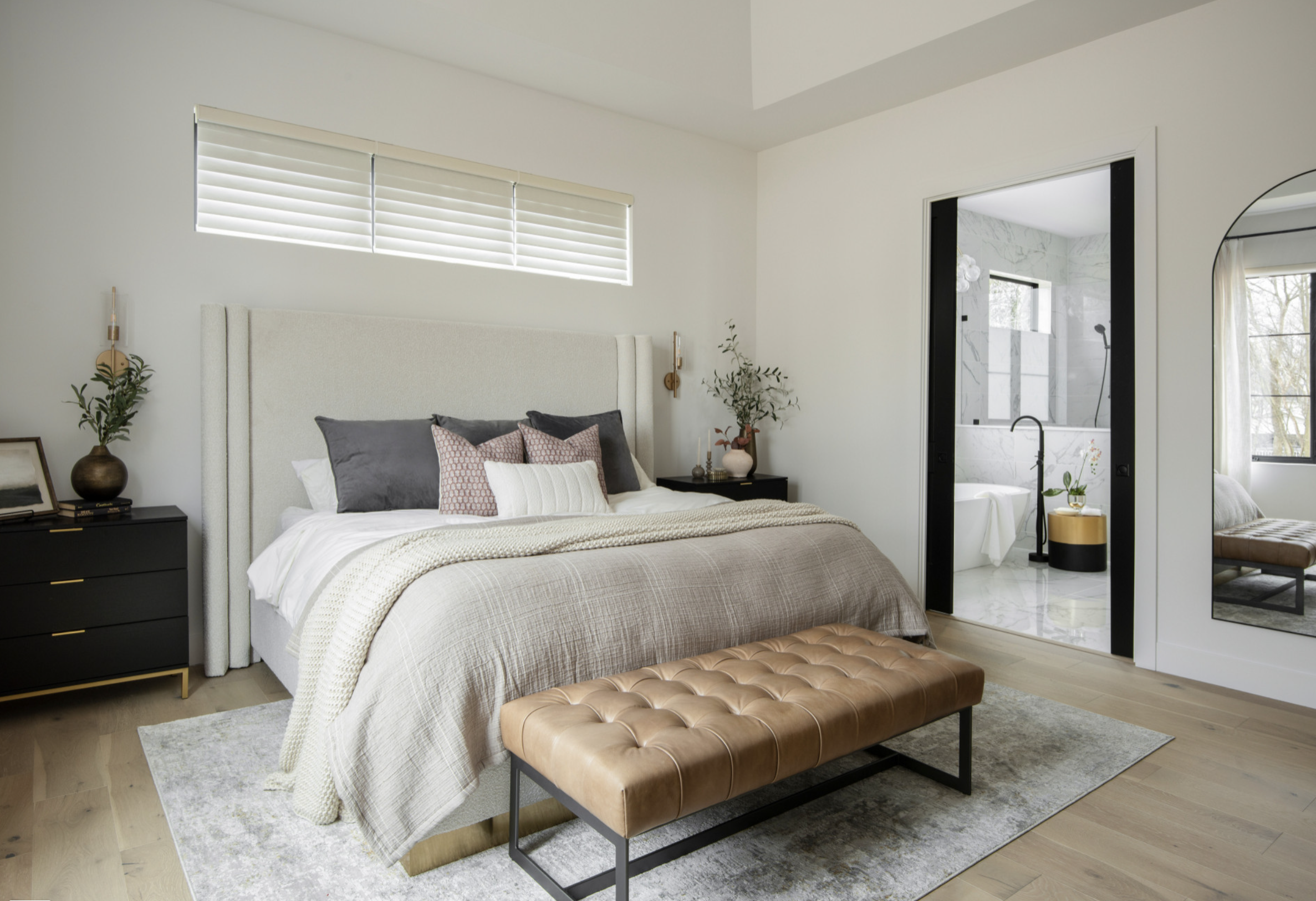 Contemporary bedroom with beige bed linens, multiple pillows, black nightstand, and a doorway leading to a bathroom with marble walls and a bathtub.