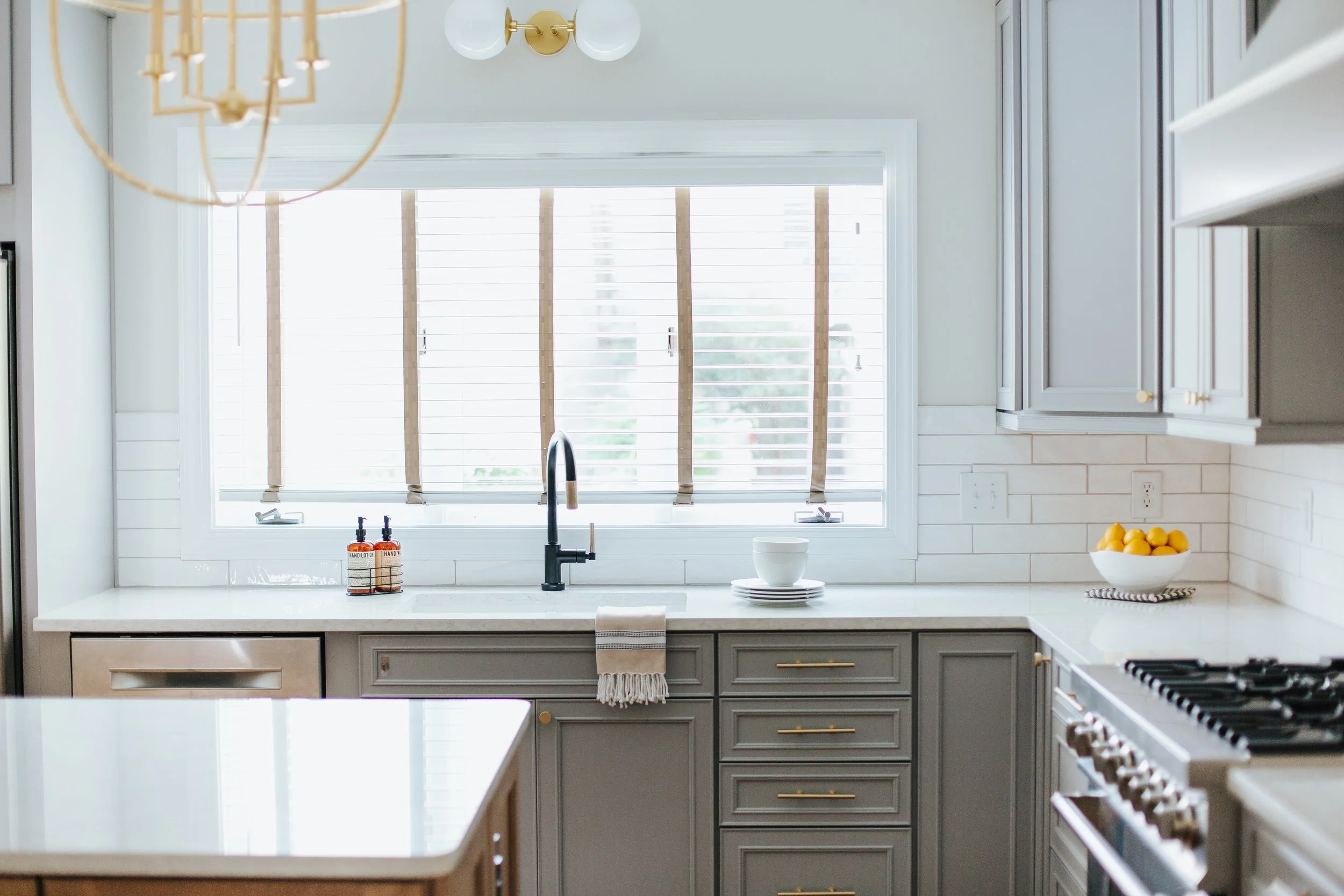 A modern kitchen with a white countertop, grey cabinets with gold handles, and a large window with white blinds. On the counter, there are two bottles of hand lotion, a white bowl, and a stack of small plates. There are lemons in a white bowl on the right side of the counter. The kitchen also has a black faucet and a stove on the right side.