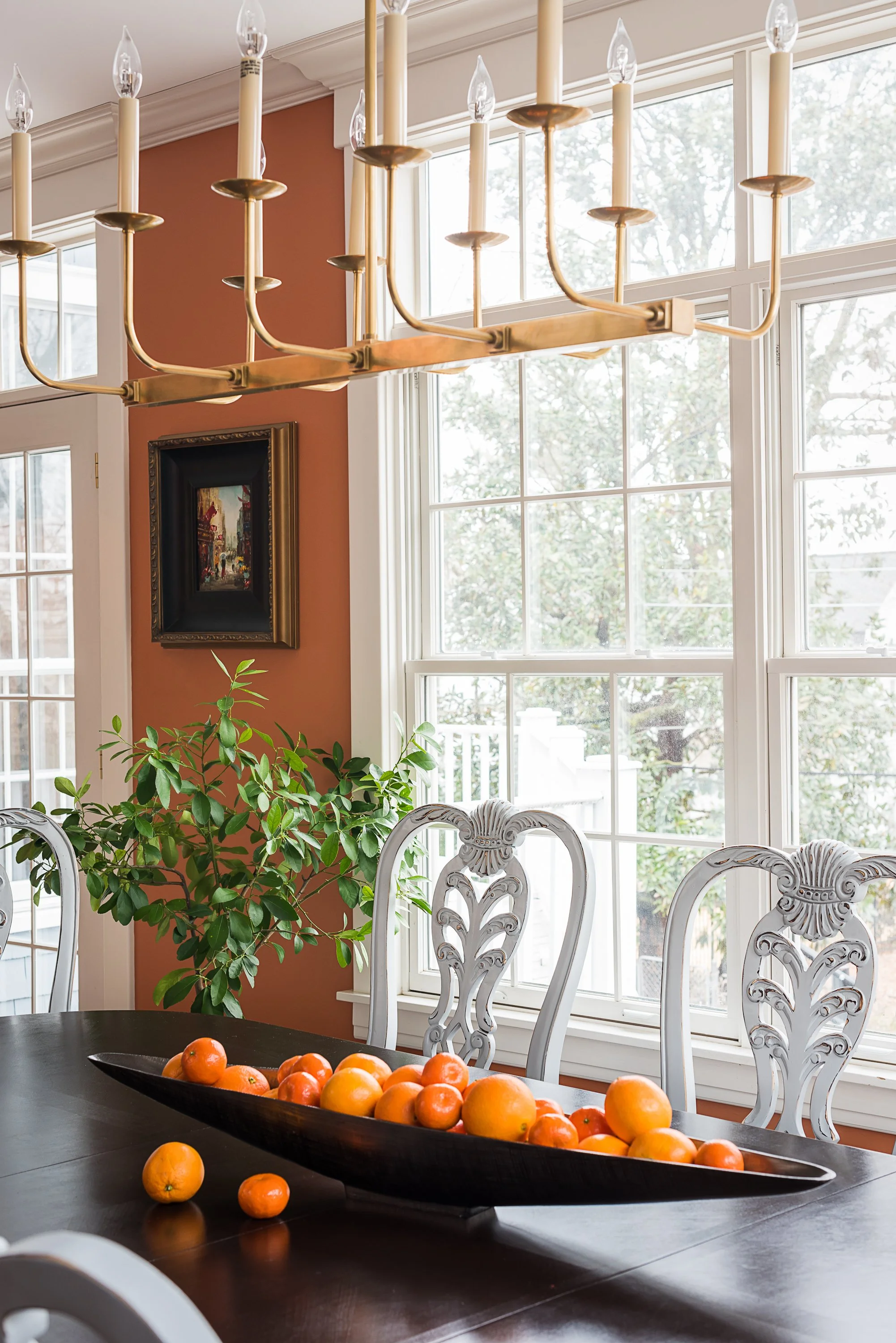 A dining room with a black oval bowl filled with oranges on a dark wooden table, surrounded by white ornate chairs. Large windows letting in natural light, a potted plant and a framed painting on a burnt orange accent wall, and a gold chandelier hanging from the ceiling.