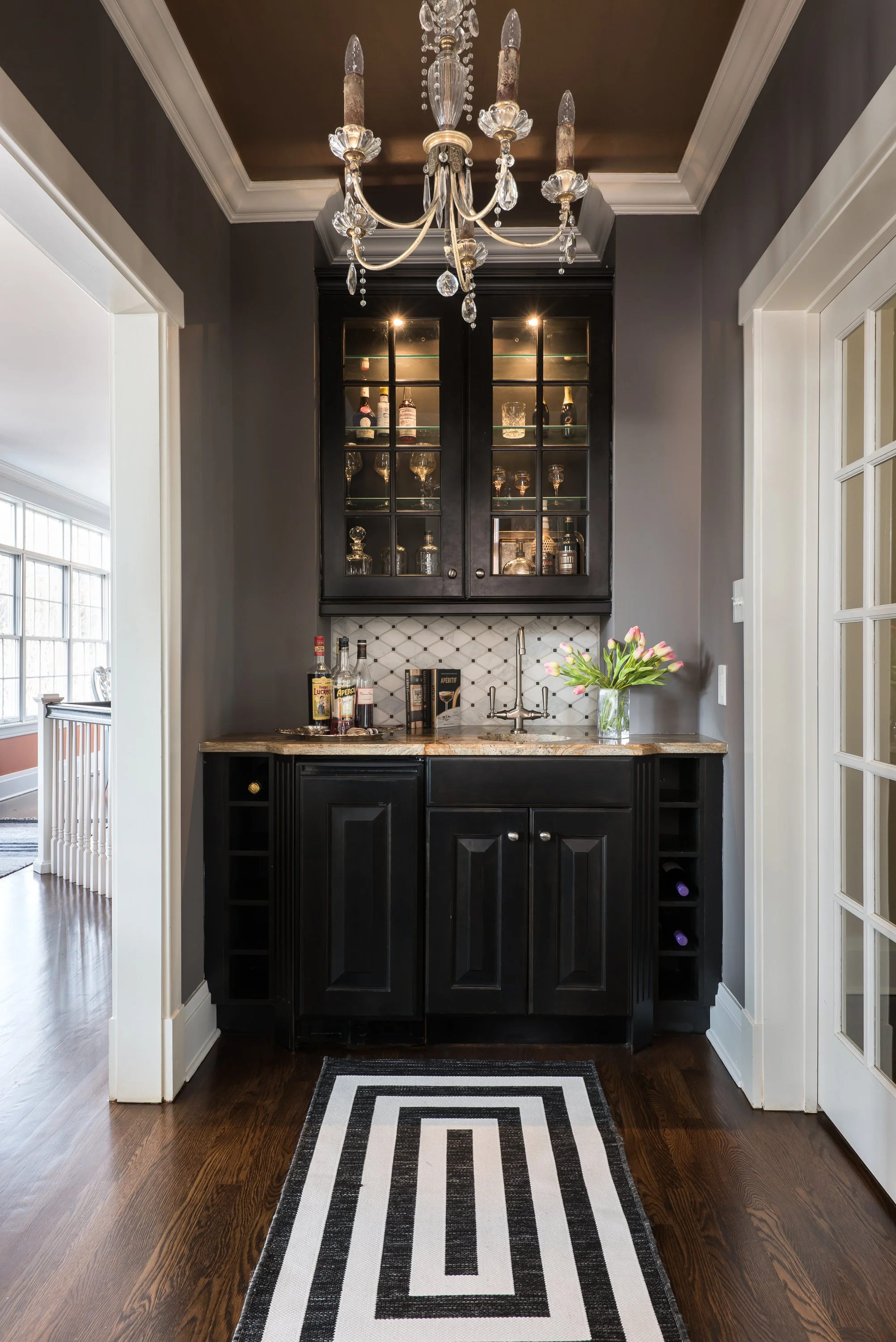 A black bar cabinet with a wine rack built in. Above the cabinet is a black wall-mounted cabinet with glass doors, displaying glasses and bottles. A chandelier with crystals hangs from the ceiling. In front of the cabinet, there is a geometric patterned rug on dark wood flooring.