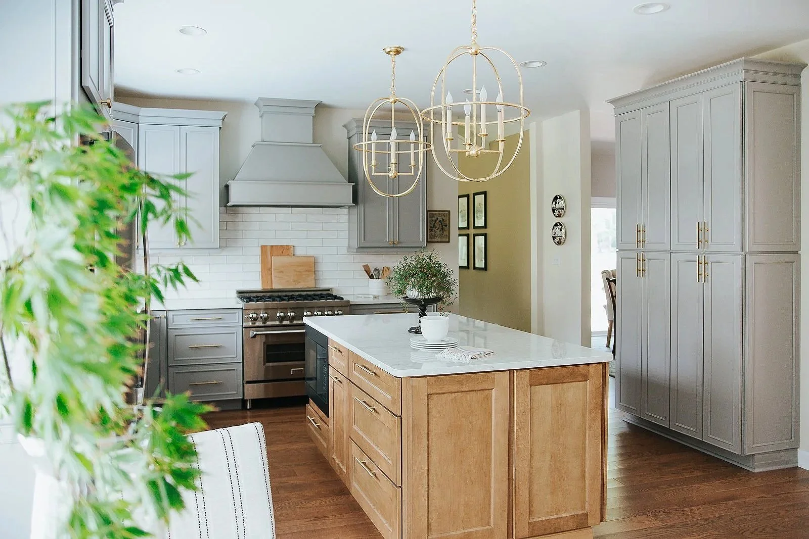 Modern kitchen with gray cabinets, a white island with a wooden base, stainless steel oven, and white subway tile backsplash. There are two gold chandeliers hanging from the ceiling, and decorative plates on the wall. Green plants are in the foreground.