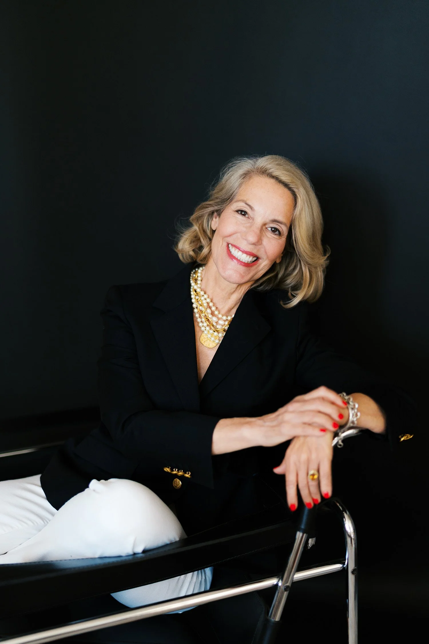 A smiling woman with blonde hair, wearing a black blazer, pearl jewelry, and red lipstick, sitting in a black chair with a dark background.