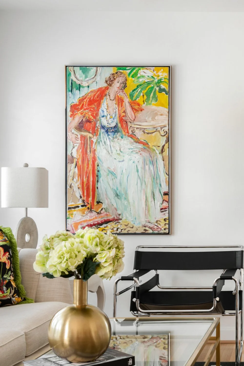 A contemporary living room moment, including a framed colorful painting of a woman, a white lamp, beige sofa with a patterned pillow, a glass-top coffee table and a black lounge chair.