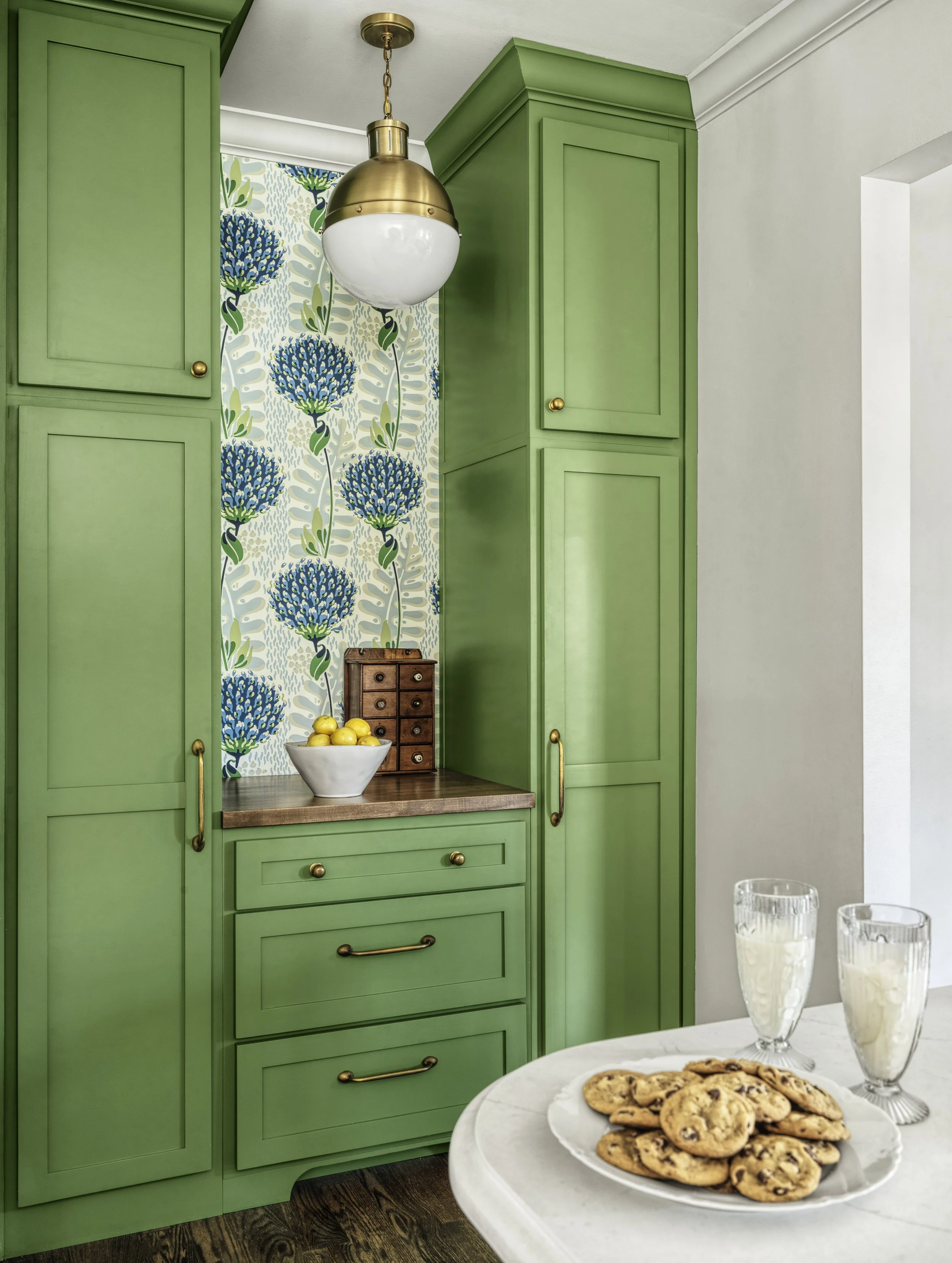 Green kitchen cabinets with brass hardware, a patterned wallpaper with blue flowers, a bowl of lemons on a counter, and a marble-topped table with cookies and milk.