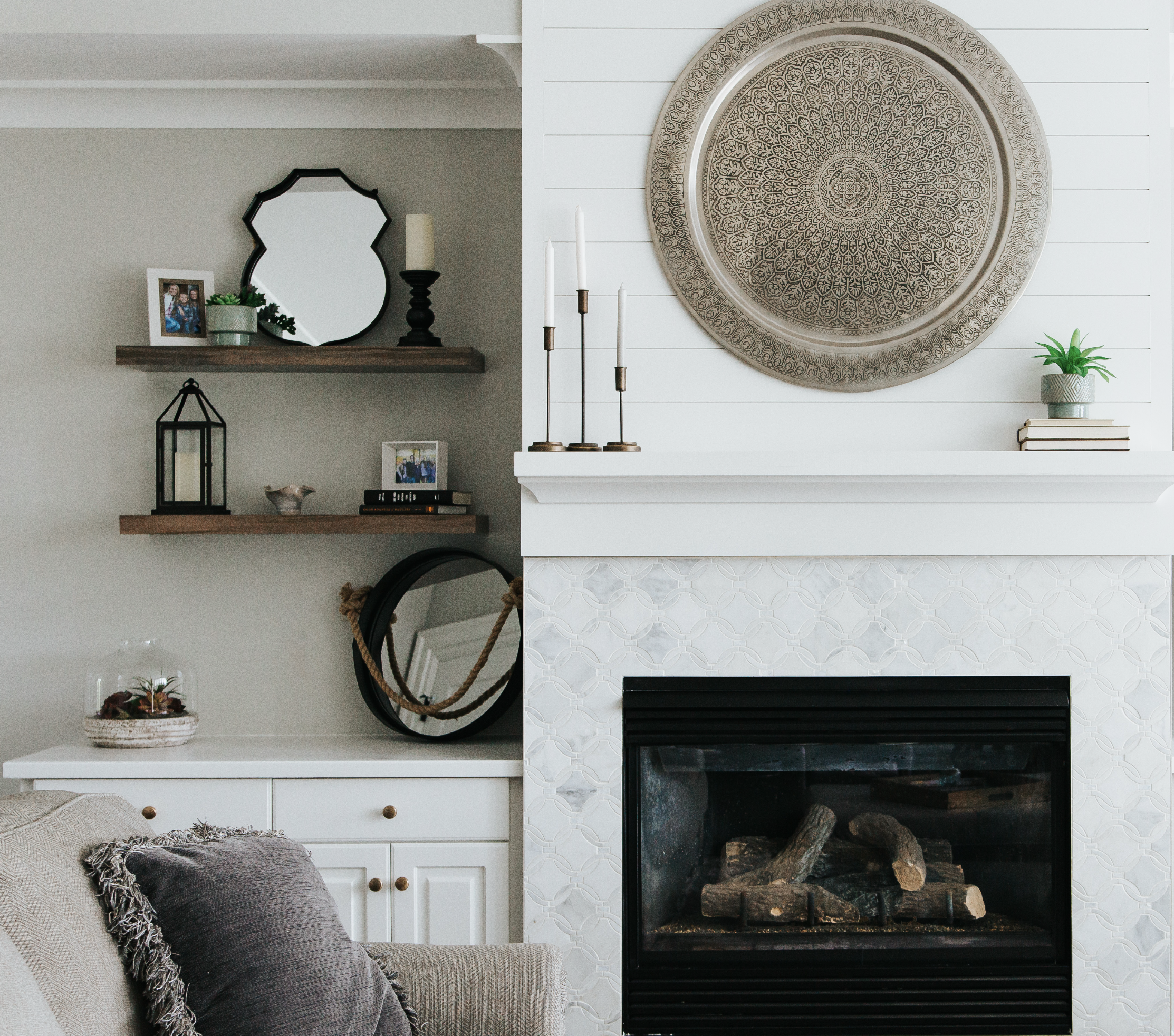 Living room fireplace with large decorative round wall art, white shiplap wall paneling, candles, potted plants, and framed family photos on shelves.