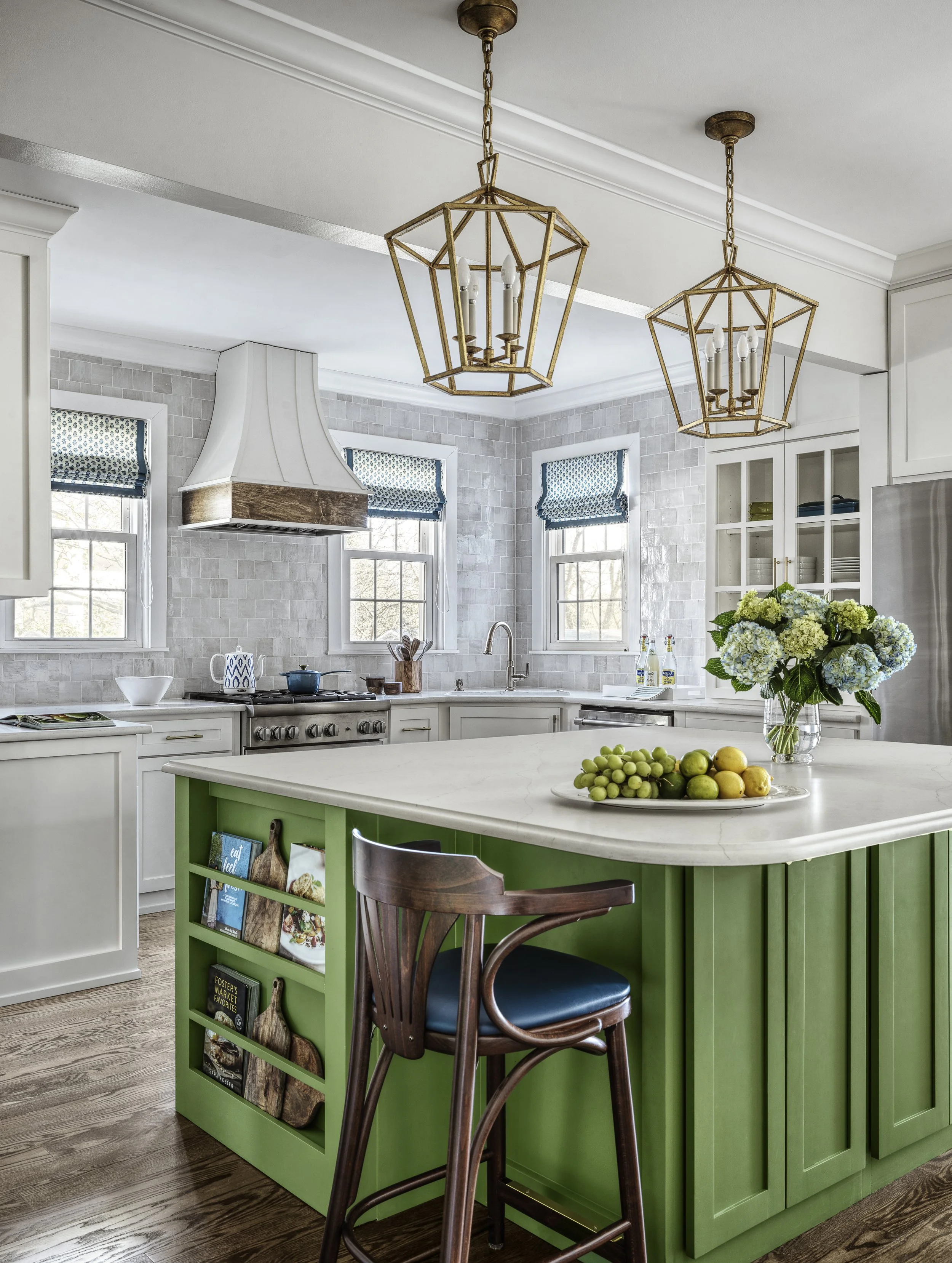 Kitchen with a green island with a magazine rack on the side and a wooden chair. Windows have blue and white patterned shades. Two gold geometric pendant lights hang from the ceiling.