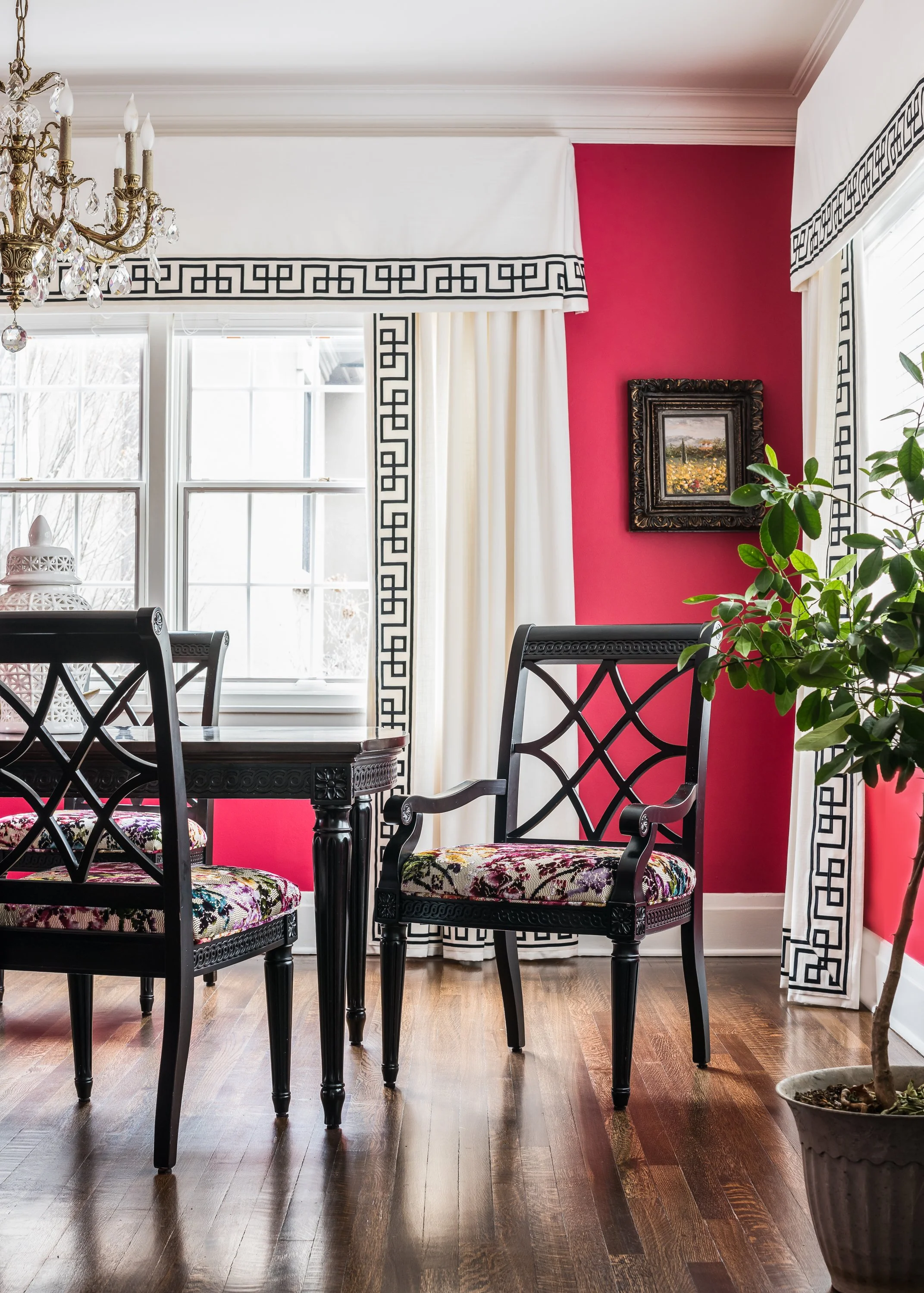 A dining room with dark wooden table and upholstered chairs, white curtains with black geometric trim, red walls, framed landscape painting, chandelier, and an indoor plant.