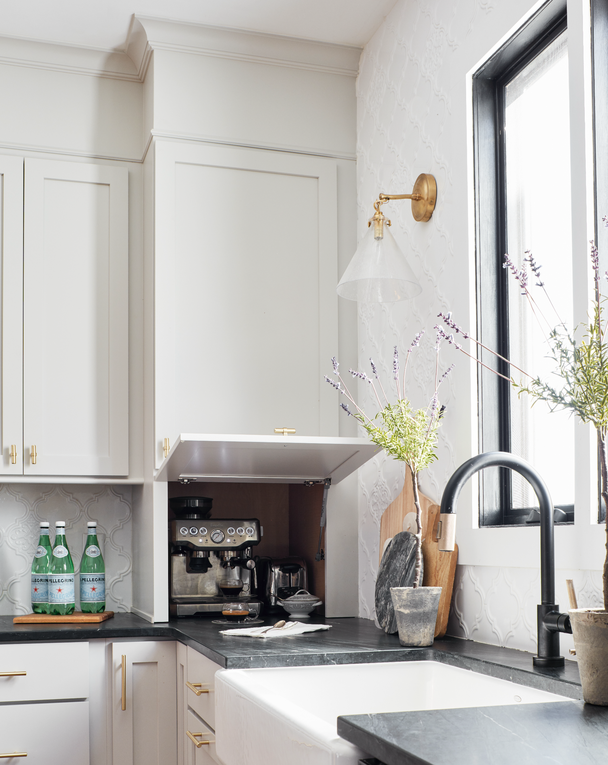 A modern kitchen with white cabinets, a black countertop, and a black window frame. There are three bottles of sparkling water on a wooden tray, an espresso machine, and some kitchen utensils on the counter. A window lets in natural light, illuminating potted plants and kitchen tools on the countertop.