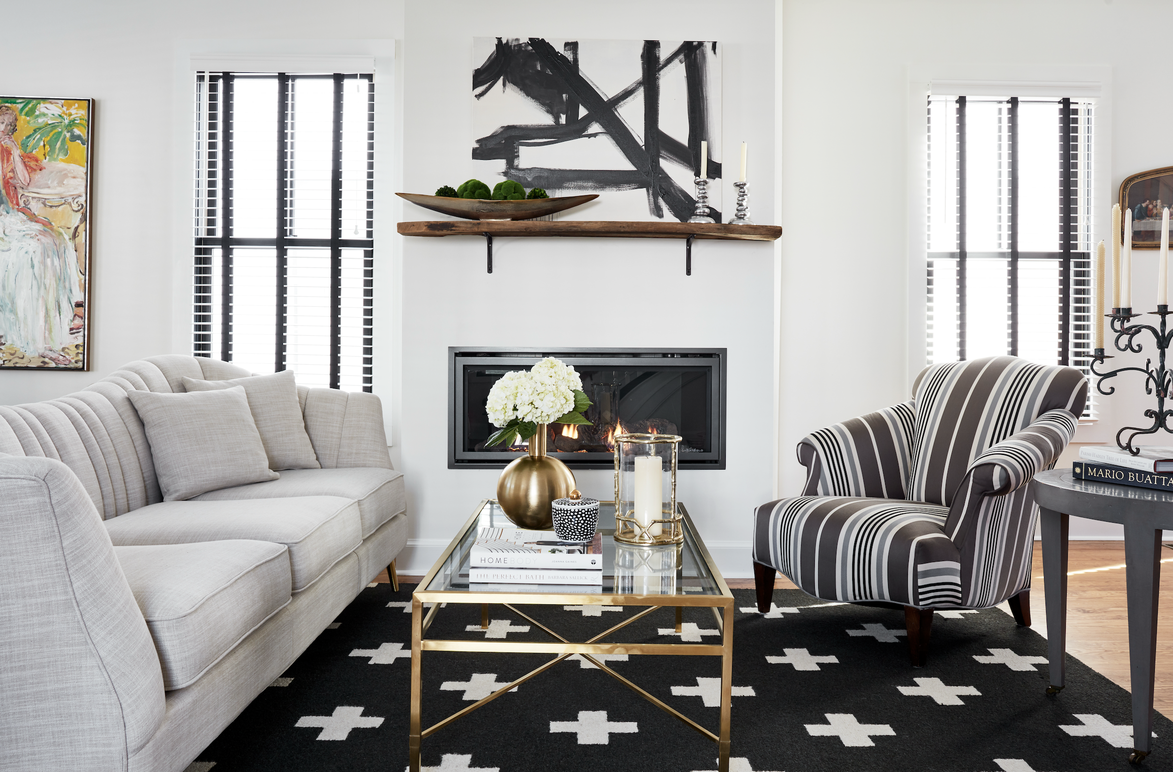 Living room with a beige sofa, black and white striped armchair, glass coffee table, fireplace, abstract black and white artwork, and decorative items including a white hydrangea bouquet and candles, with large windows and black-and-white patterned rug.