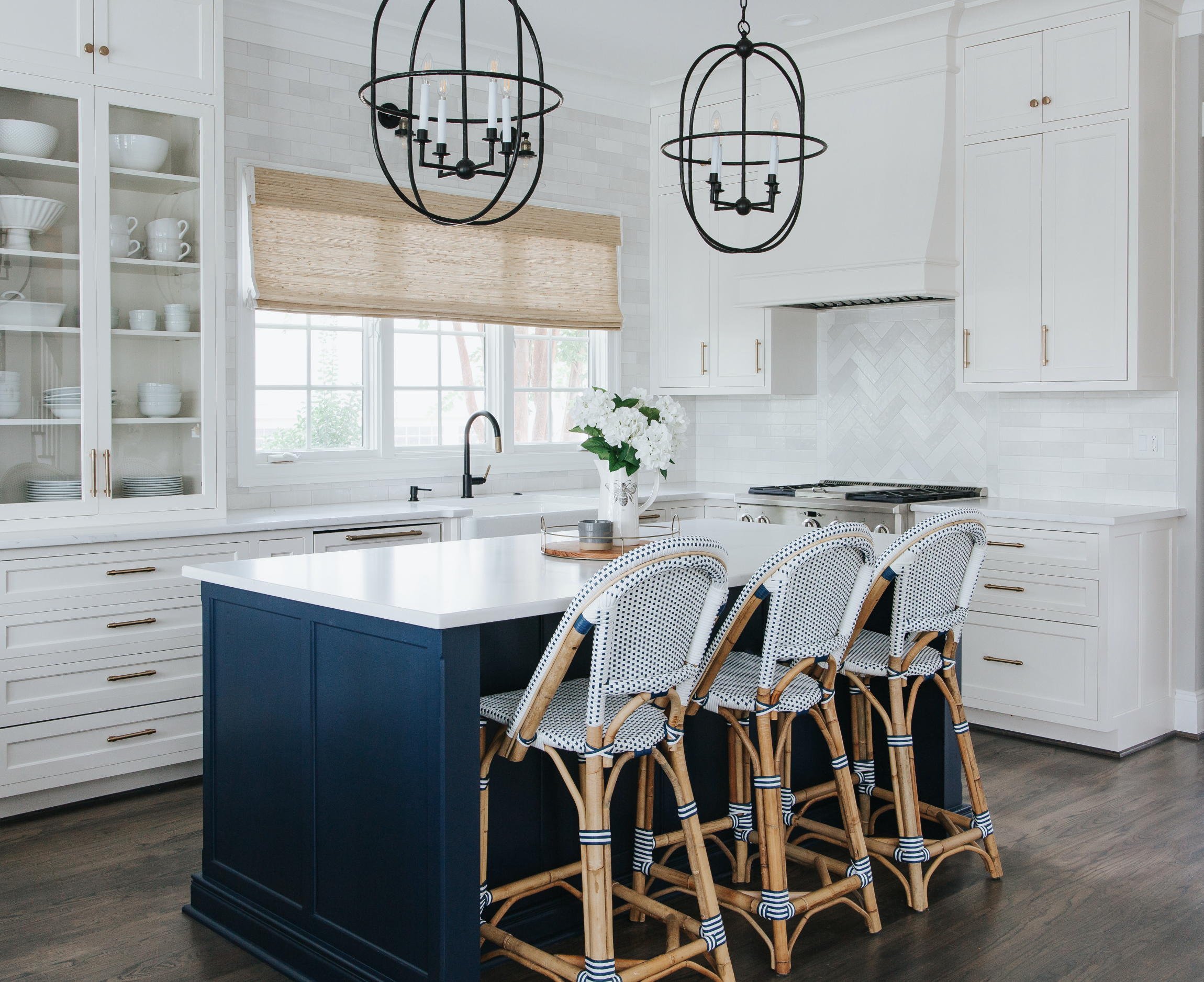 A modern kitchen with white cabinets, a navy blue island, a white countertop, a window with a woven shade and black light fixtures.
