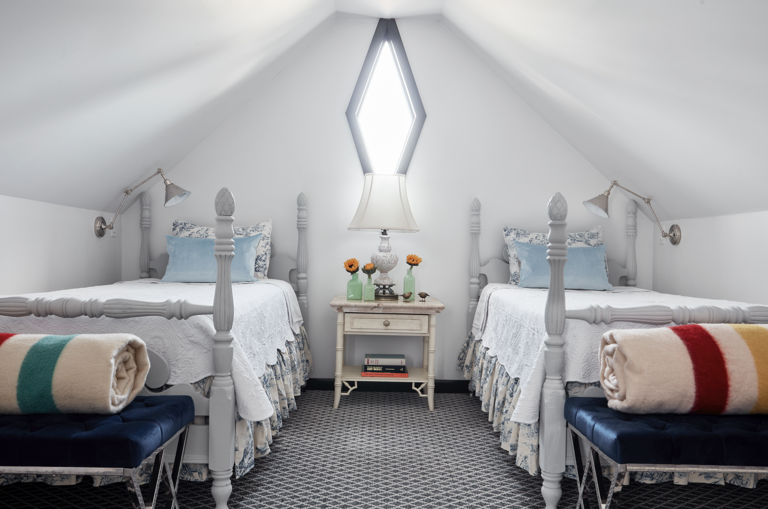 A bedroom with two white four-poster beds with blue and white patterned bedding and pillows. A nightstand with a white lamp and small orange flowers in green vases is between the beds. There are two adjustable wall-mounted reading lamps above each bed, and a diamond-shaped window on the sloped ceiling. The floor is covered with a black and white patterned rug.
