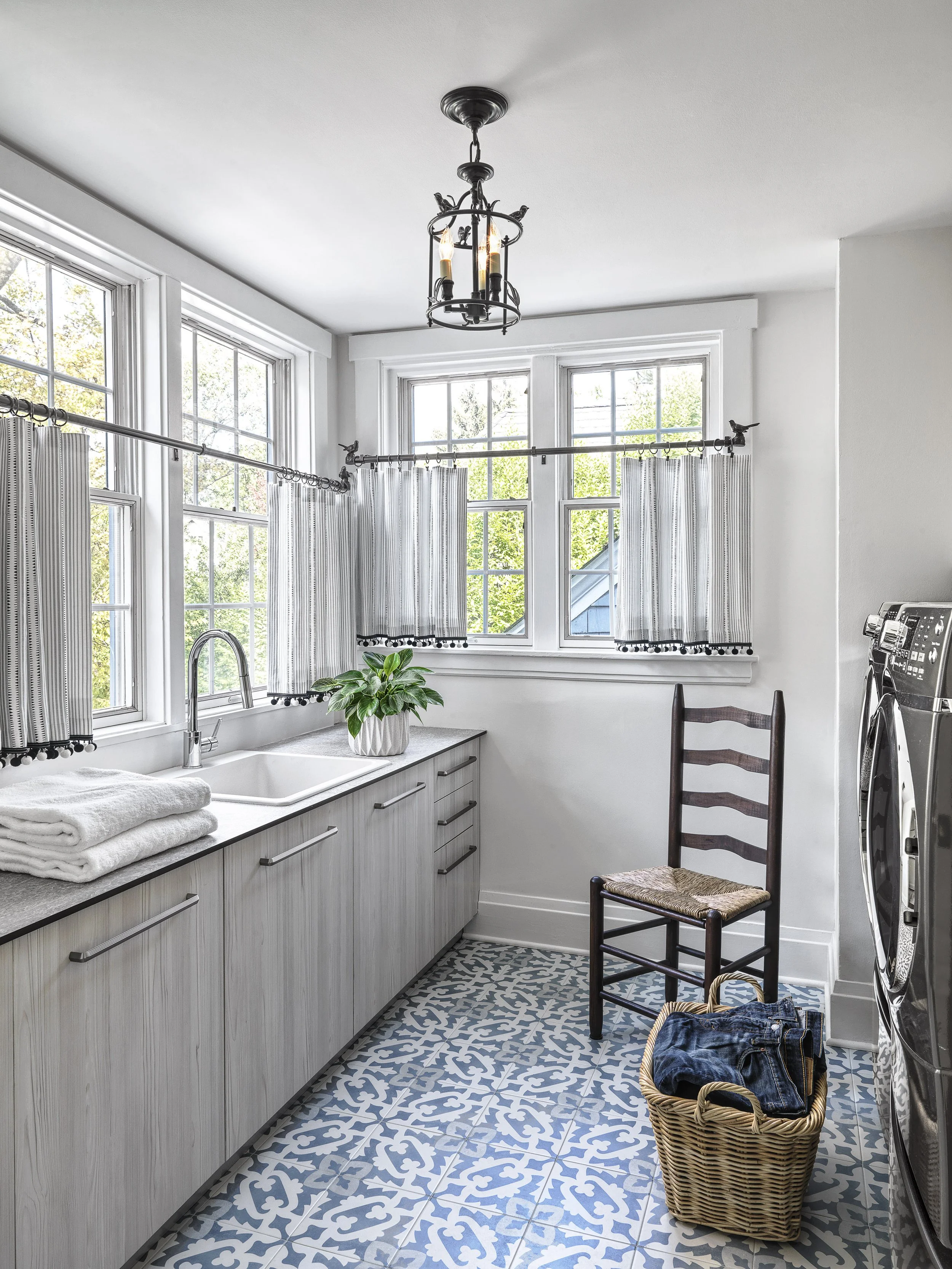 A bright laundry room with large windows, white cabinets, a sink, a chair, a laundry basket filled with clothes, a washing machine, a potted plant, and a decorative blue and white patterned tile floor. A black chandelier hangs from the ceiling.